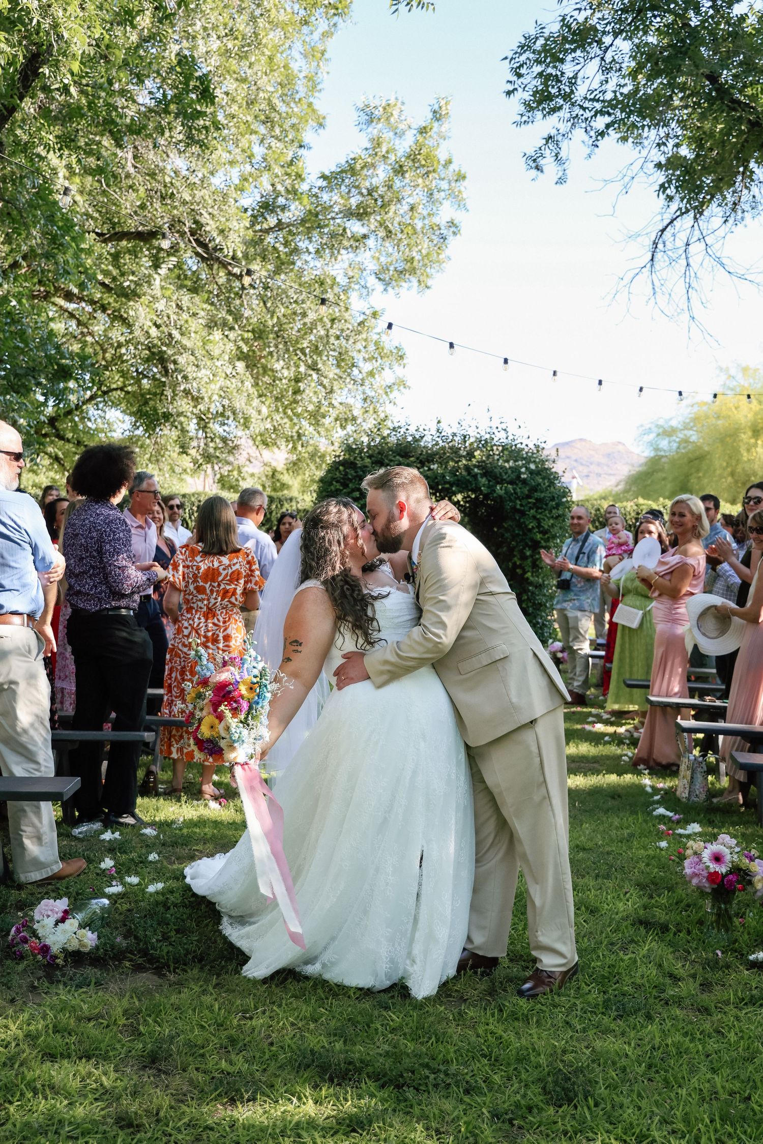 Bride and groom share a kiss at outdoor wedding ceremony on green lawn with guests and string lights.