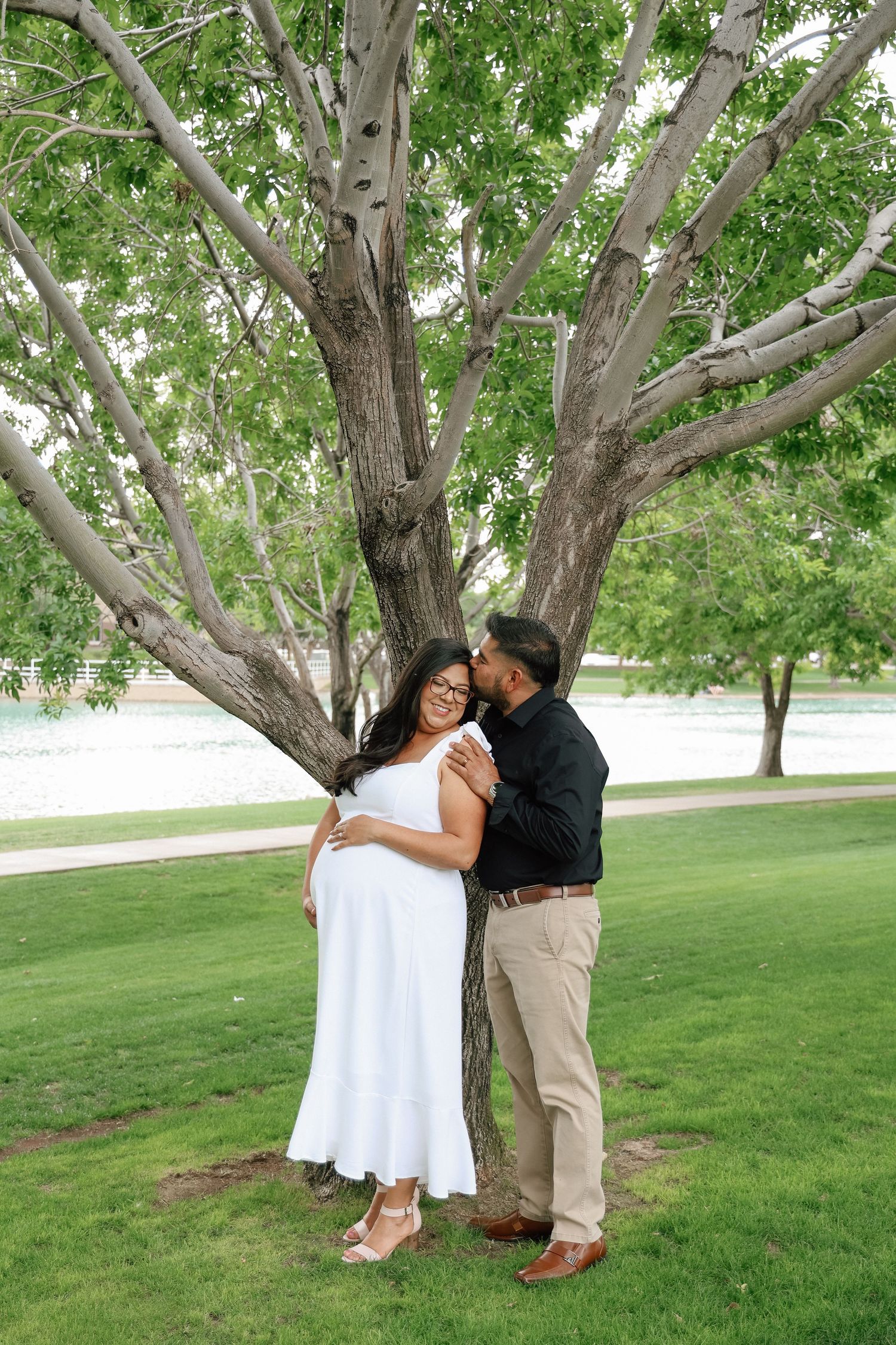 Pregnant woman in white dress leaning against tree while man in black shirt kisses her cheek in a lush park.