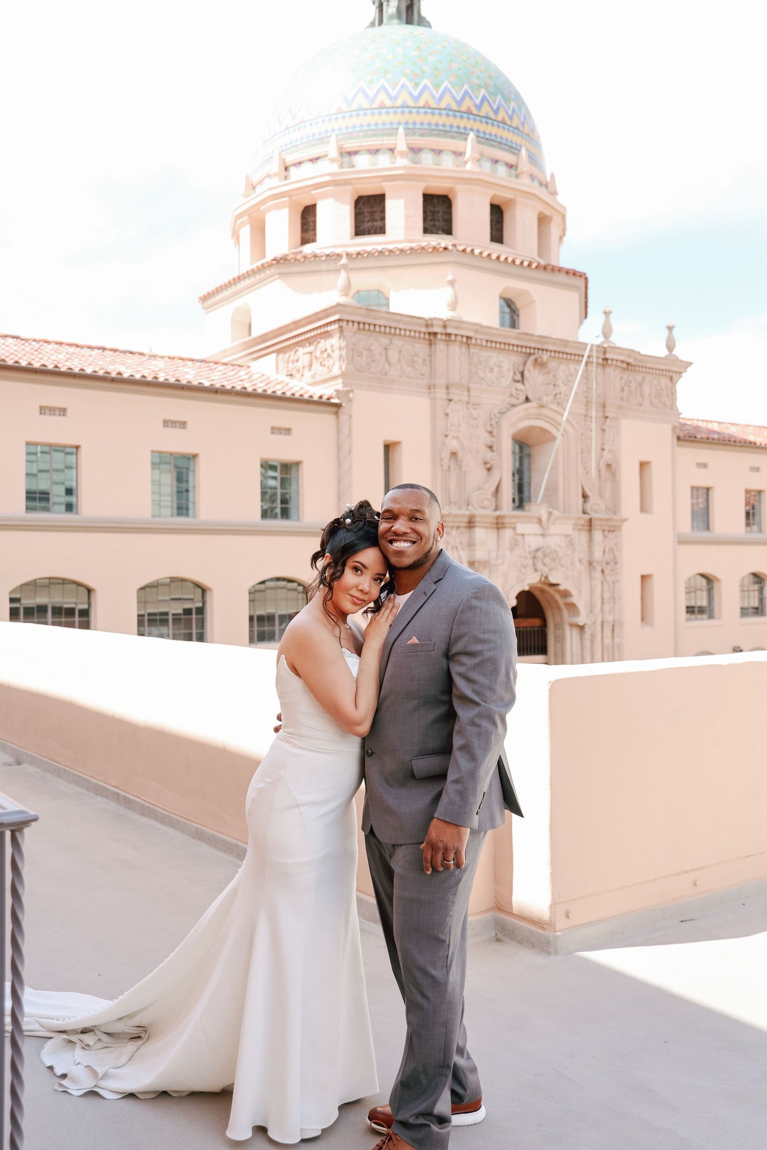 A bride and groom pose on a rooftop terrace in front of a grand building with a teal dome on their wedding day.