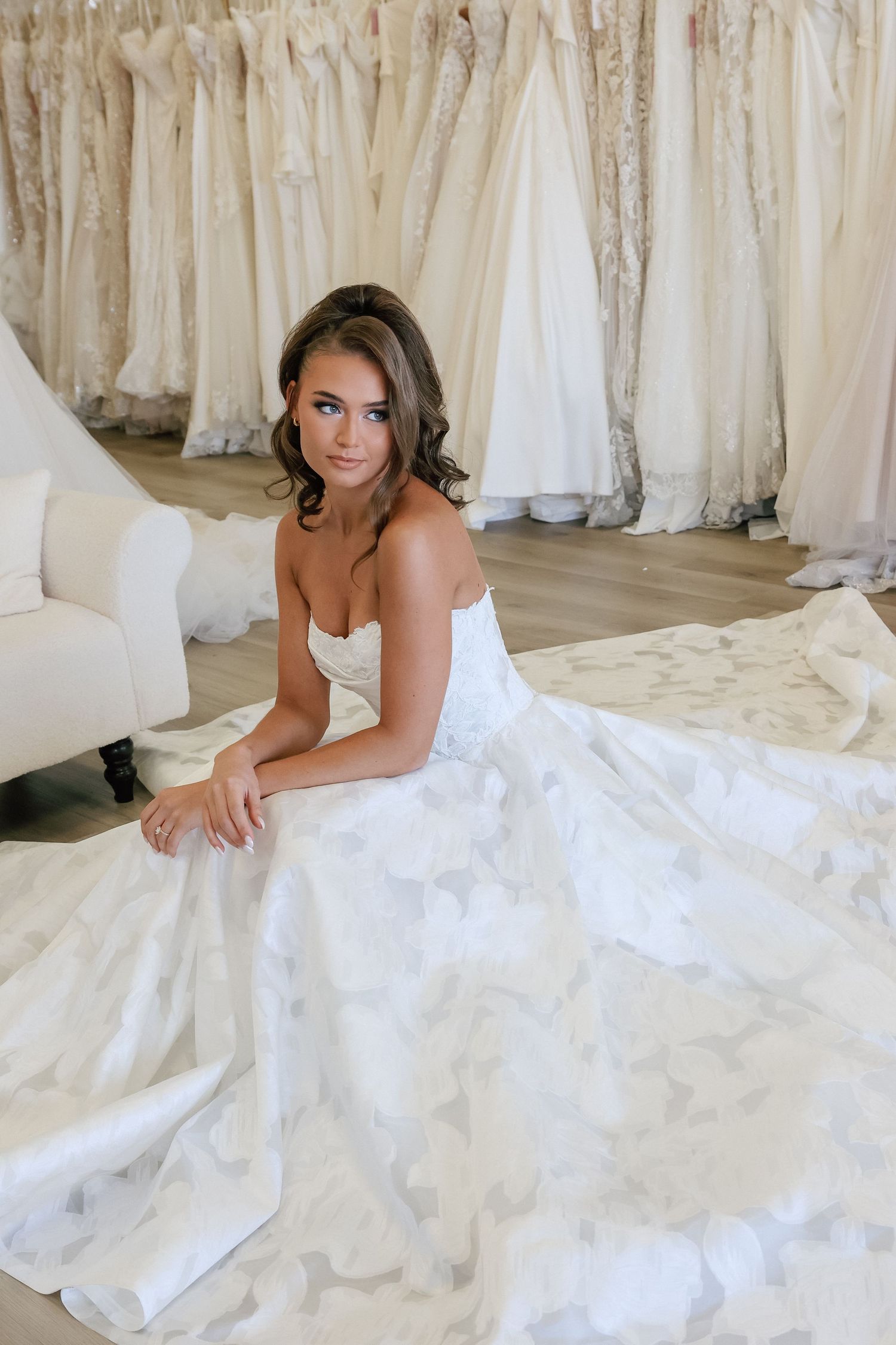 Bride in strapless white ballgown sits on floor of bridal boutique surrounded by wedding dresses on display.