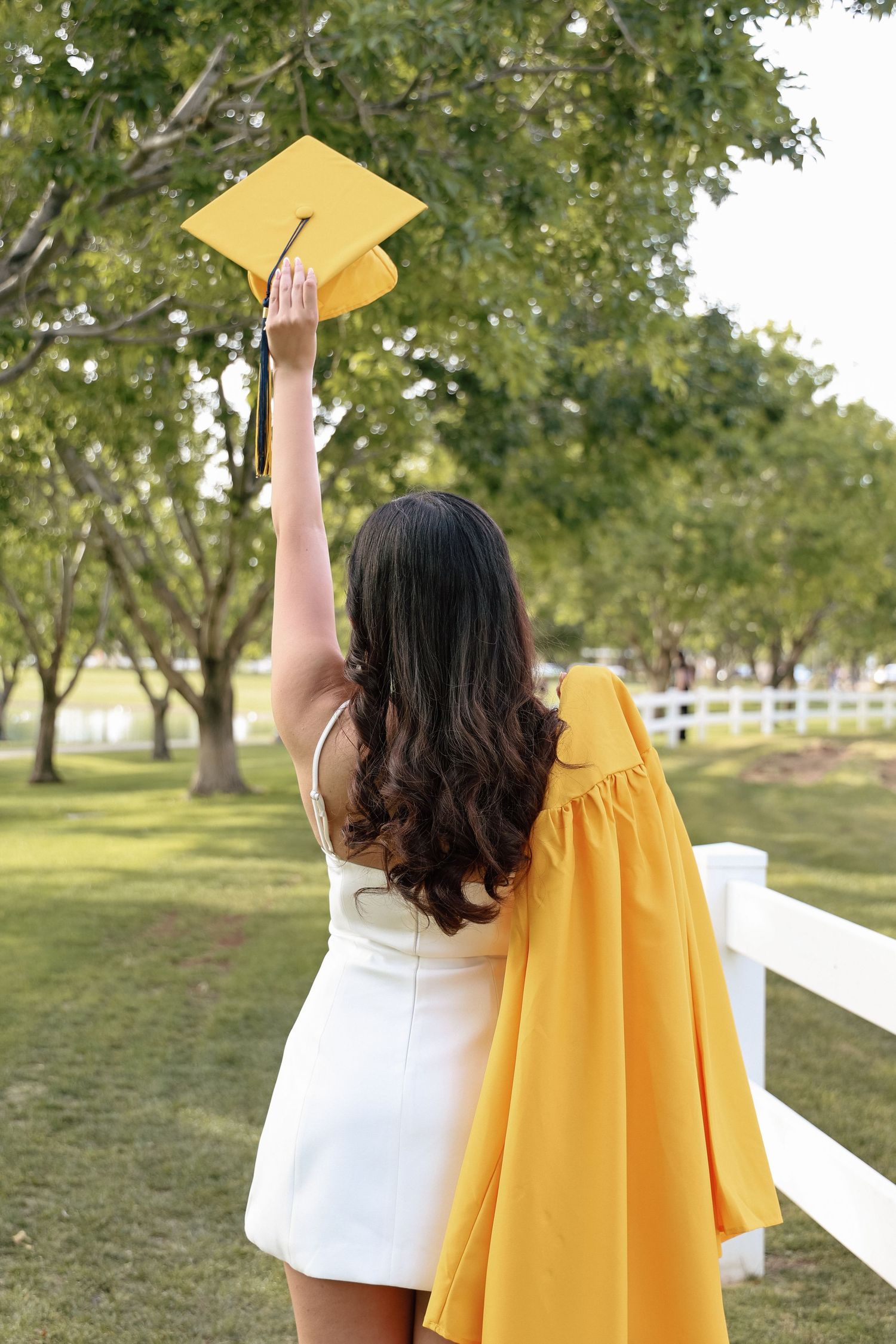 A graduate in a white dress raises her yellow cap and gown in celebration outdoors near a white fence.