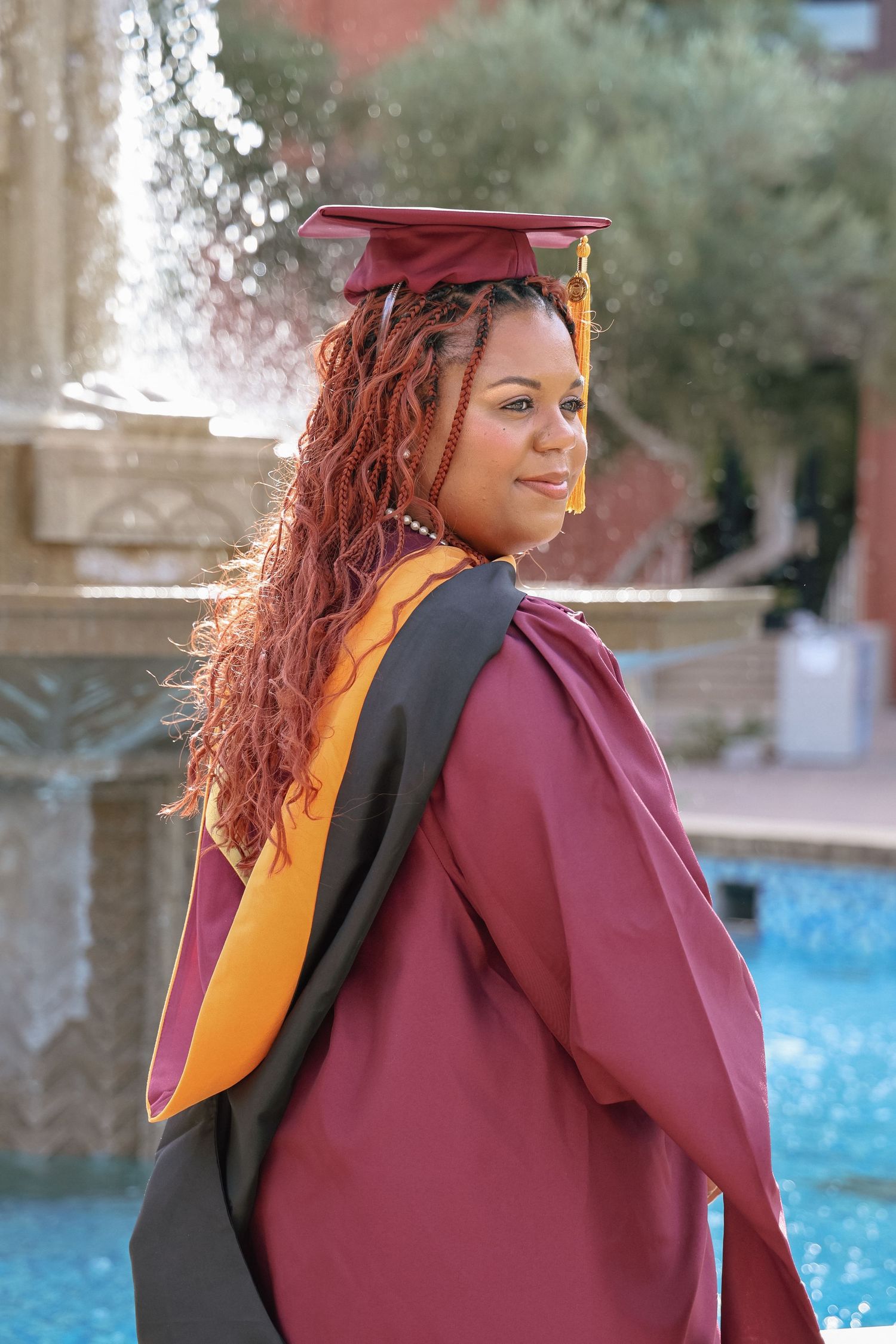 Young woman in maroon graduation gown and cap smiles over shoulder near fountain on sunny day.