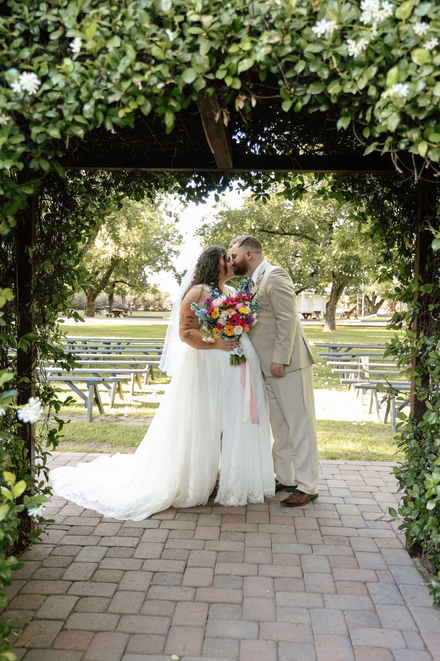 Bride and groom share first kiss under ivy-covered arbor with colorful bridal bouquet at outdoor wedding.