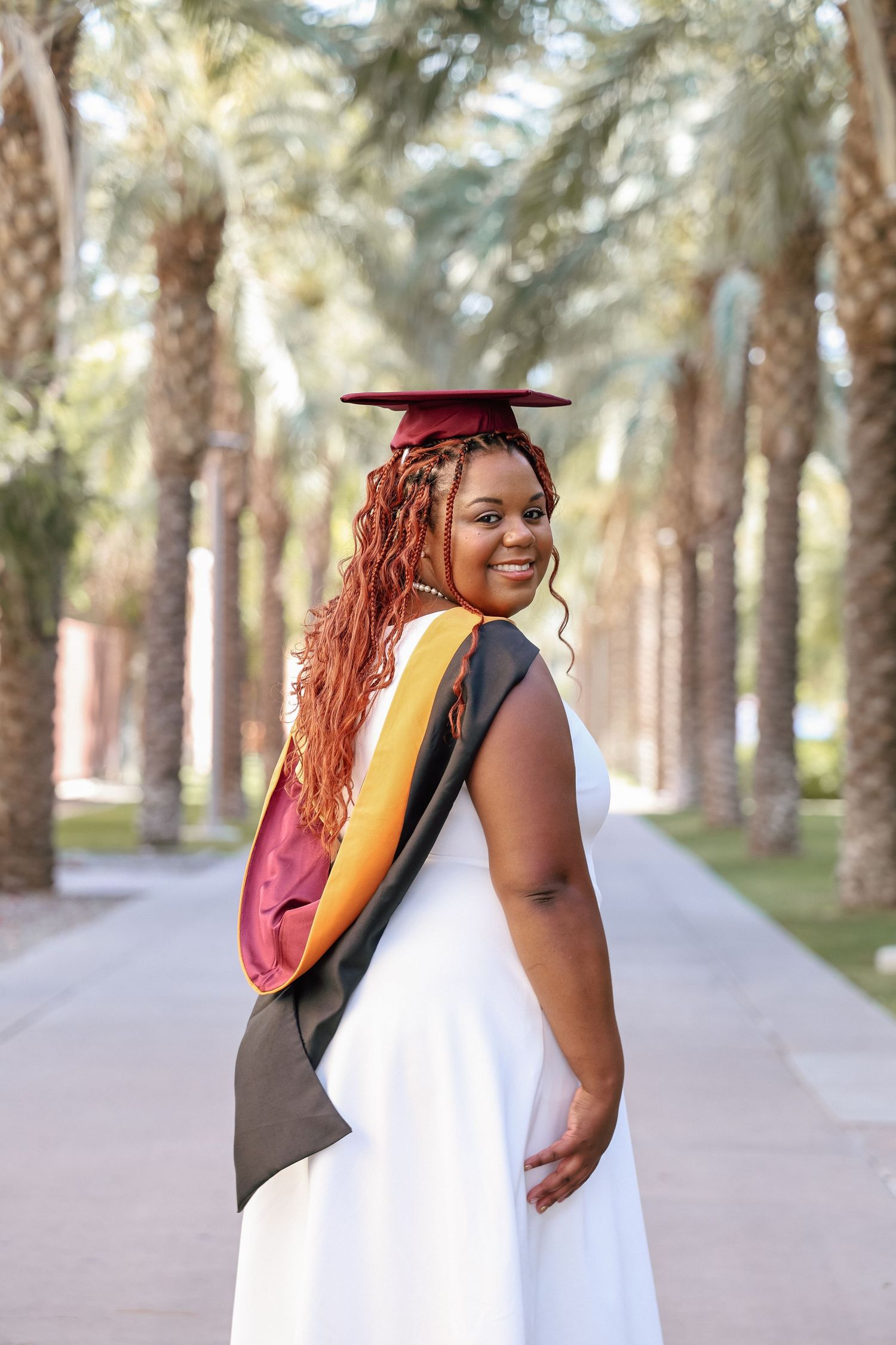 Young woman in white dress and maroon graduation cap and stole smiles outdoors among palm trees.