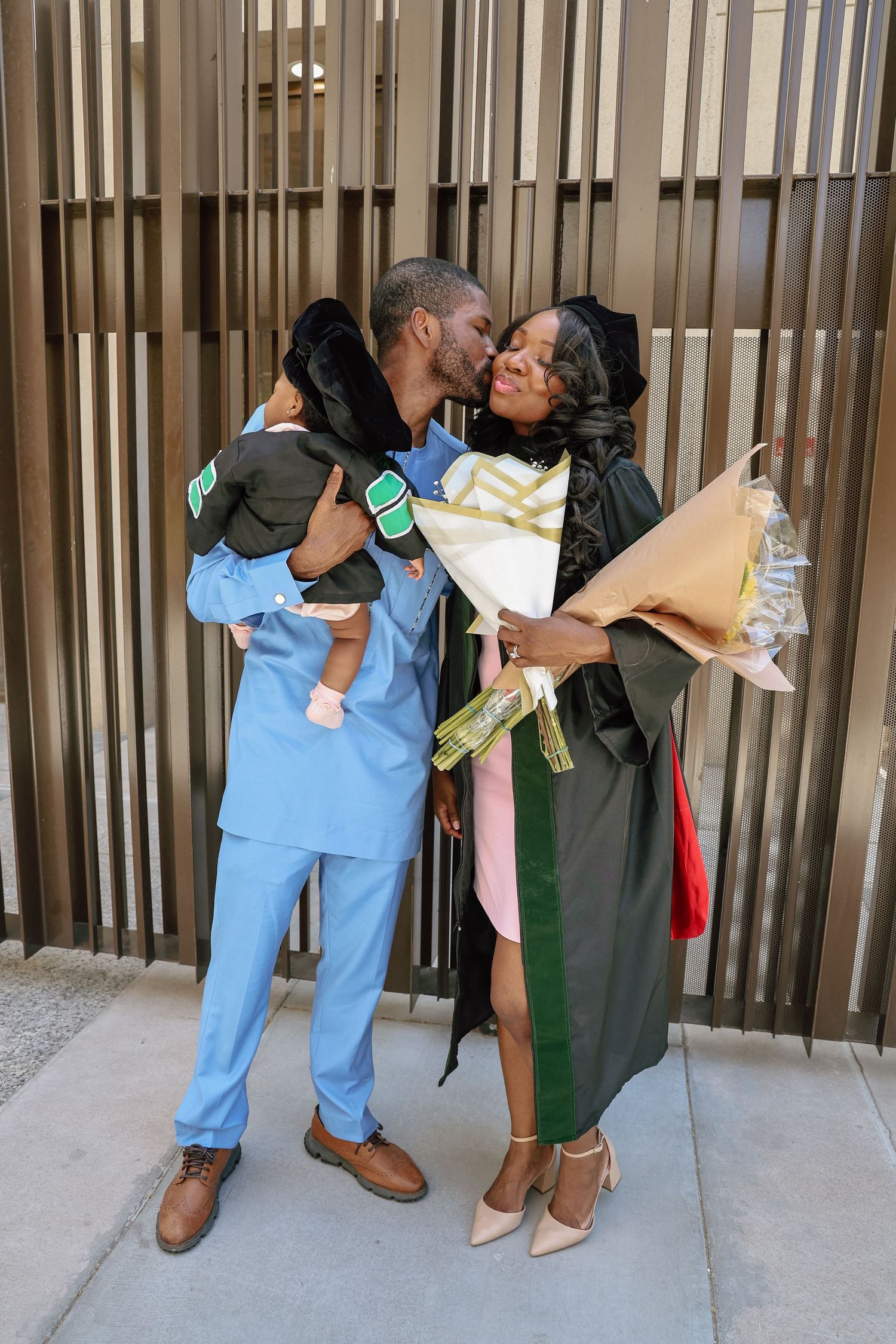 A couple in graduation gown and scrubs kissing while holding a baby and bouquet of flowers outdoors.