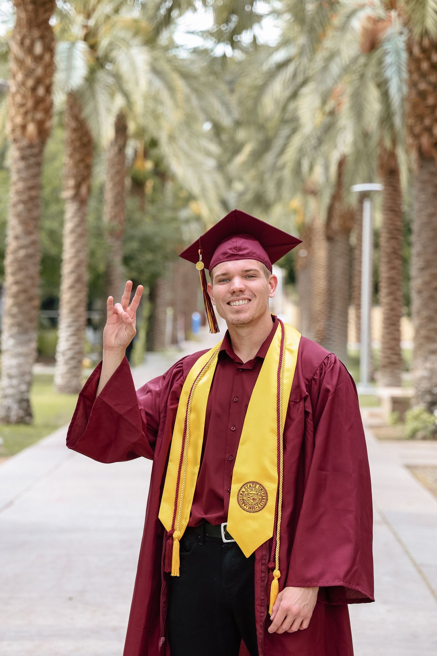 Graduate in maroon cap and gown with gold stole flashing peace sign outdoors near palm trees.