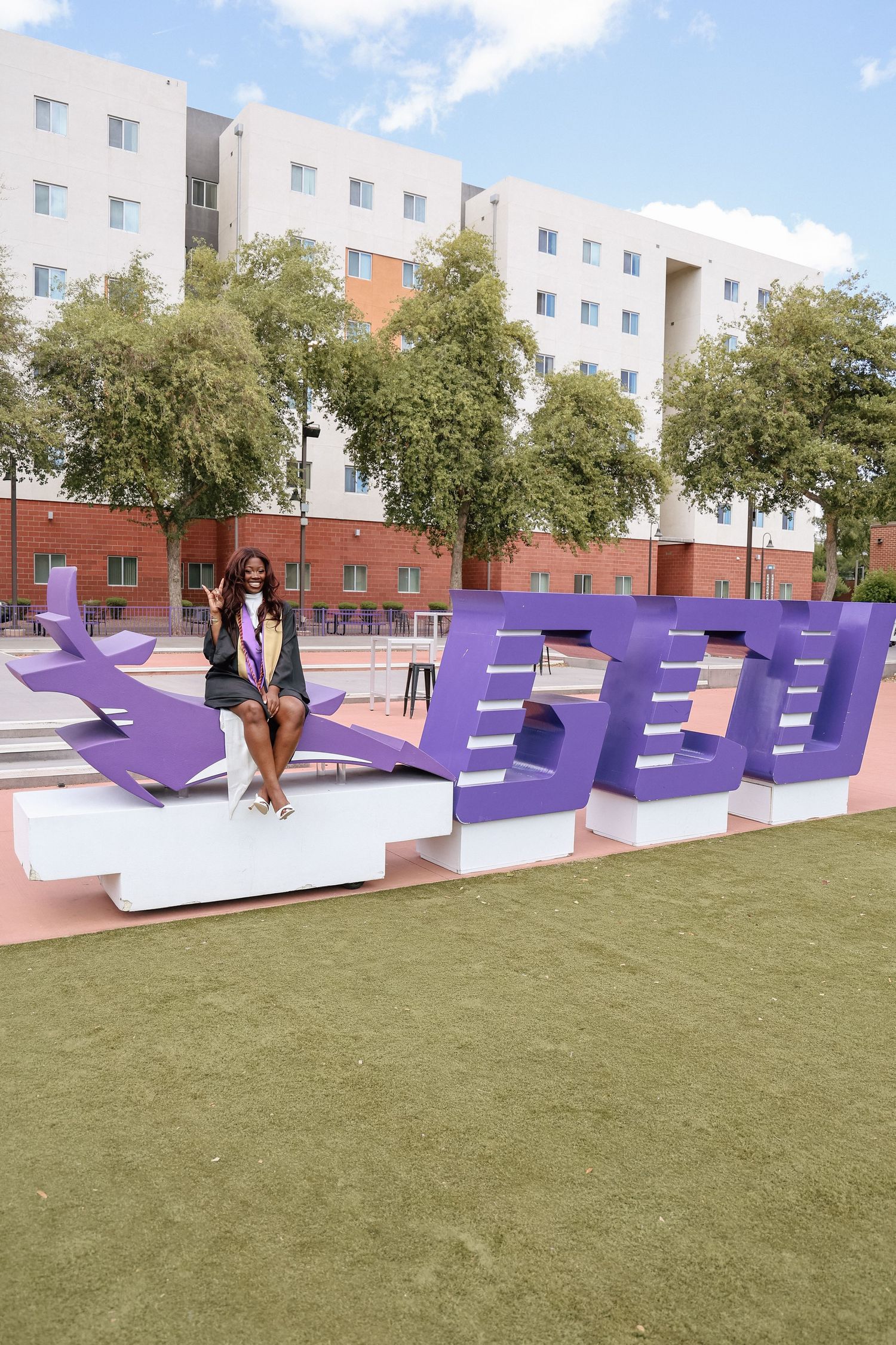 A graduate in cap and gown sits on large purple GCU letters on a sunny campus with trees and buildings in the background.