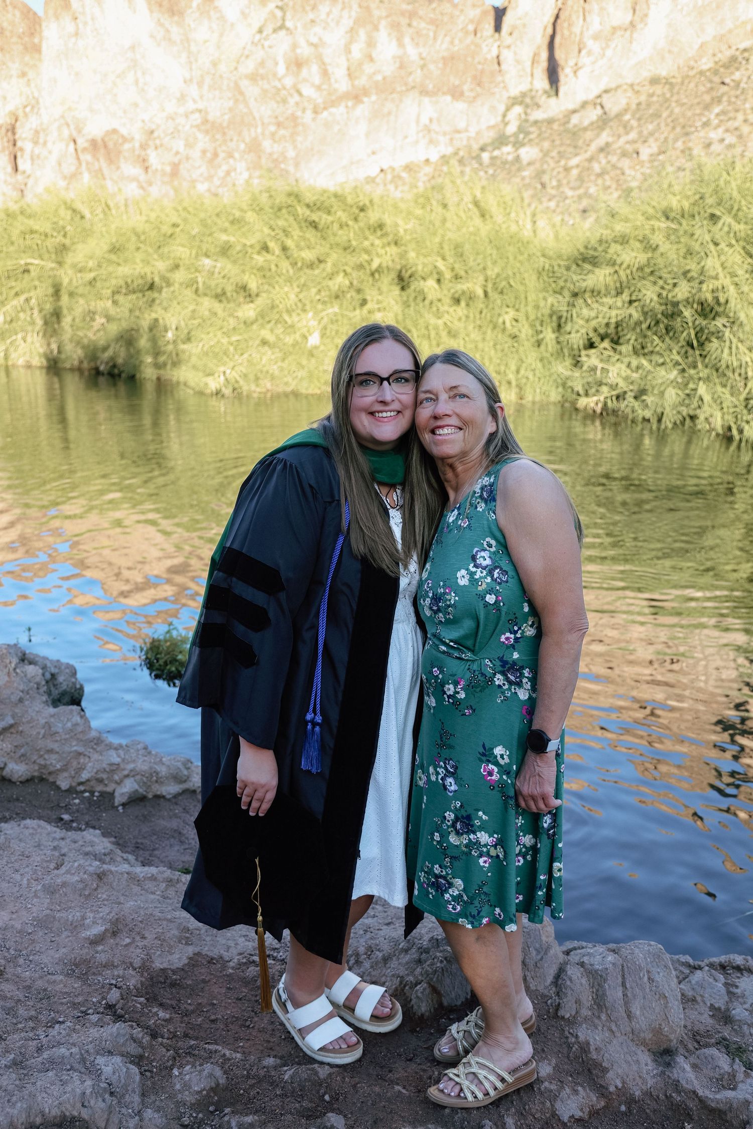 Two women pose together outdoors near a river, one wearing graduation robes and the other in a floral dress.