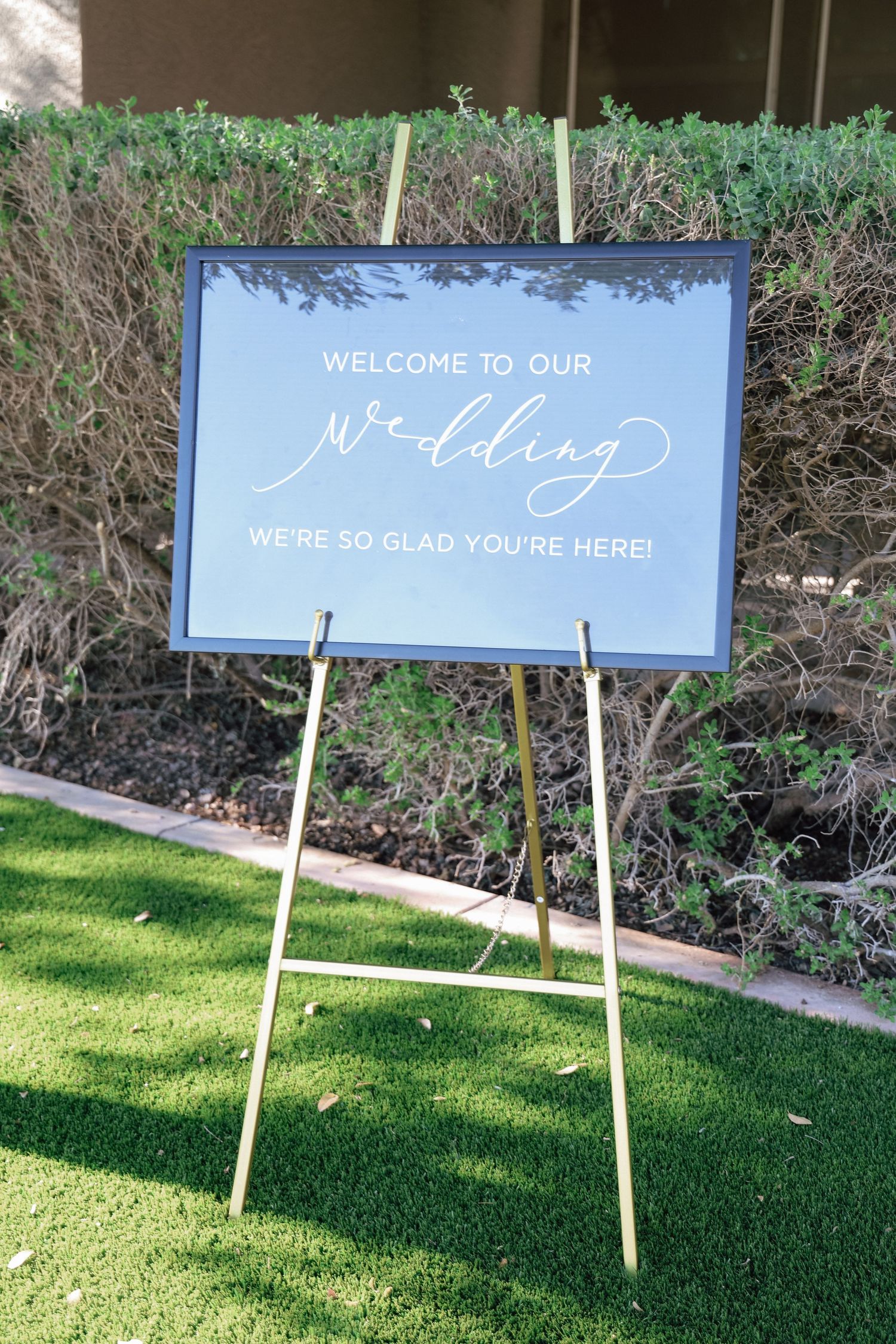 Blue wedding welcome sign on gold easel reads Welcome to Our Wedding, We're So Glad You're Here on green lawn.
