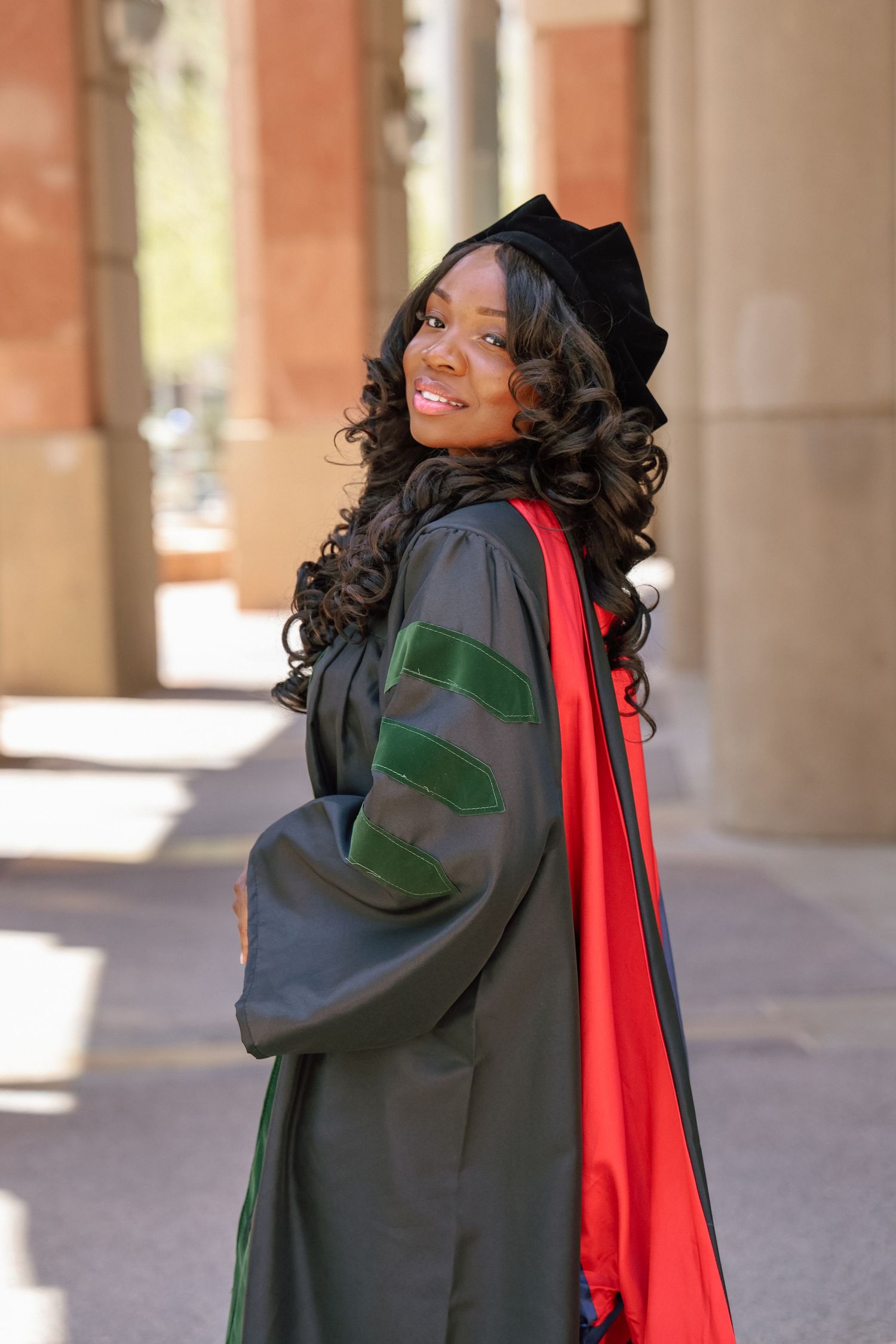 Smiling woman in black doctoral graduation gown with green velvet stripes, red hood, and mortarboard cap outdoors.