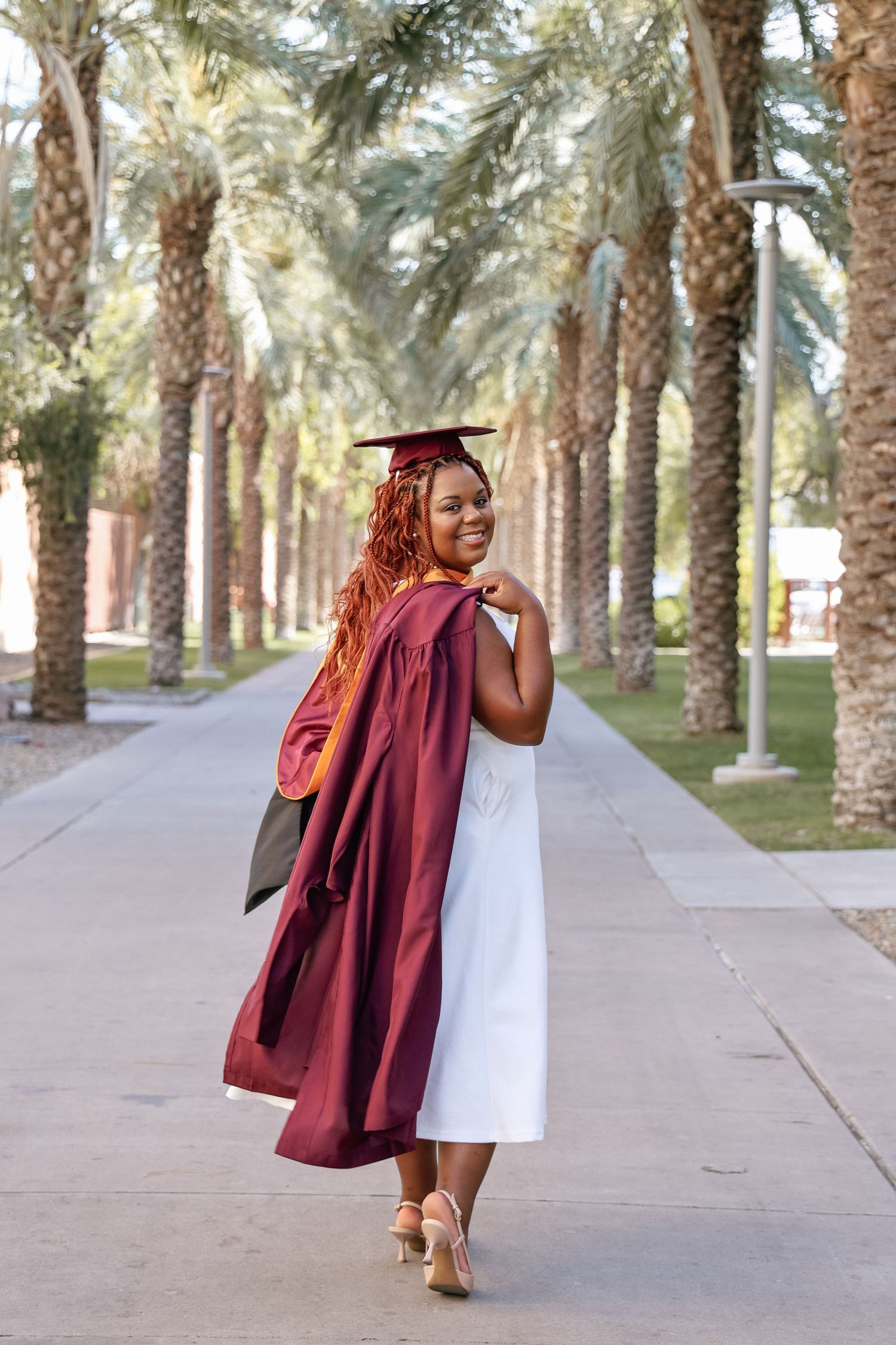 Smiling graduate in maroon cap and gown walks among palm trees, looking back over her shoulder on a sunny day.