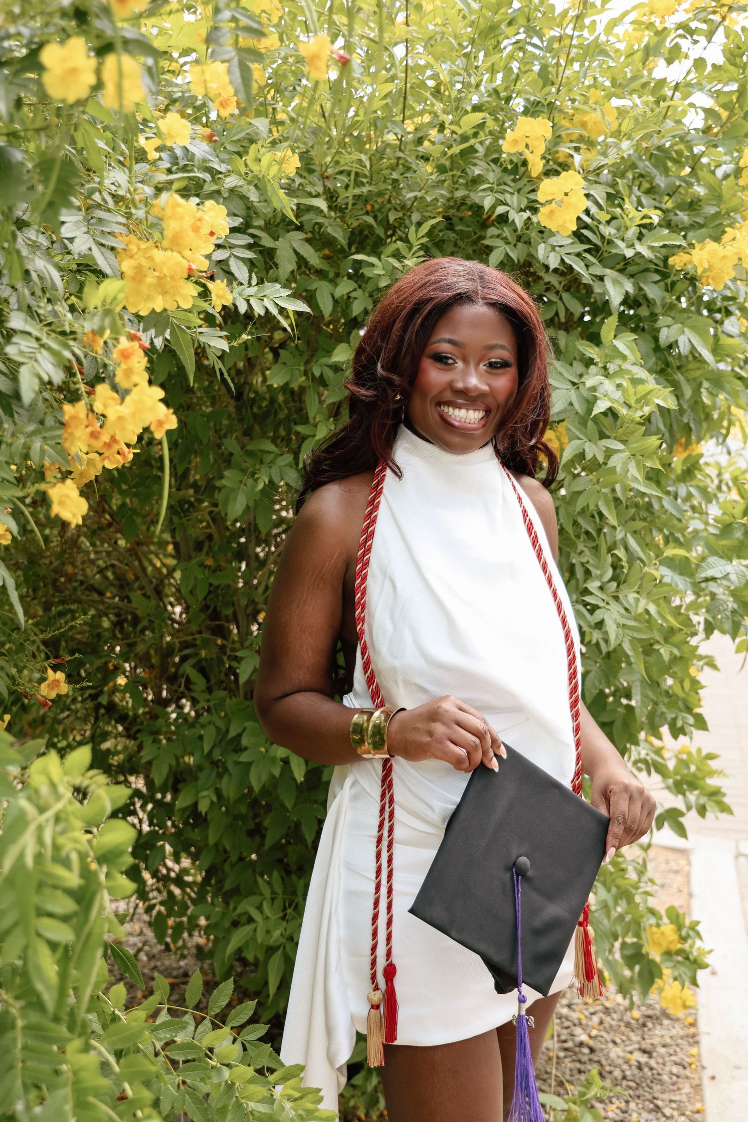 Smiling graduate in white dress holding cap with red honor cords, posing outdoors near yellow flowering bushes.