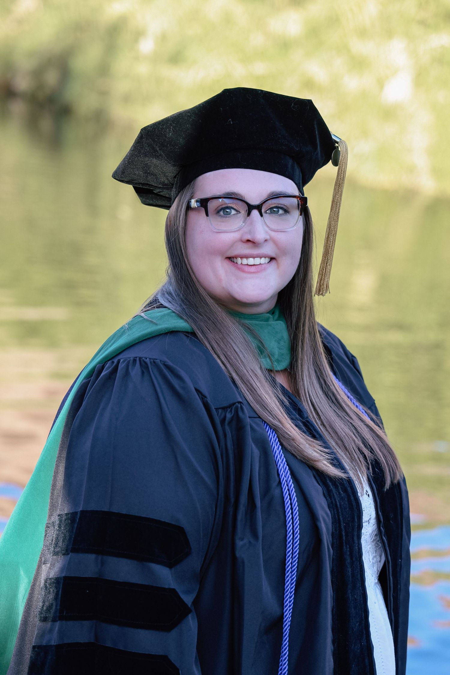 Smiling woman in black doctoral graduation gown and cap with gold tassel and green hood posing outdoors near water.