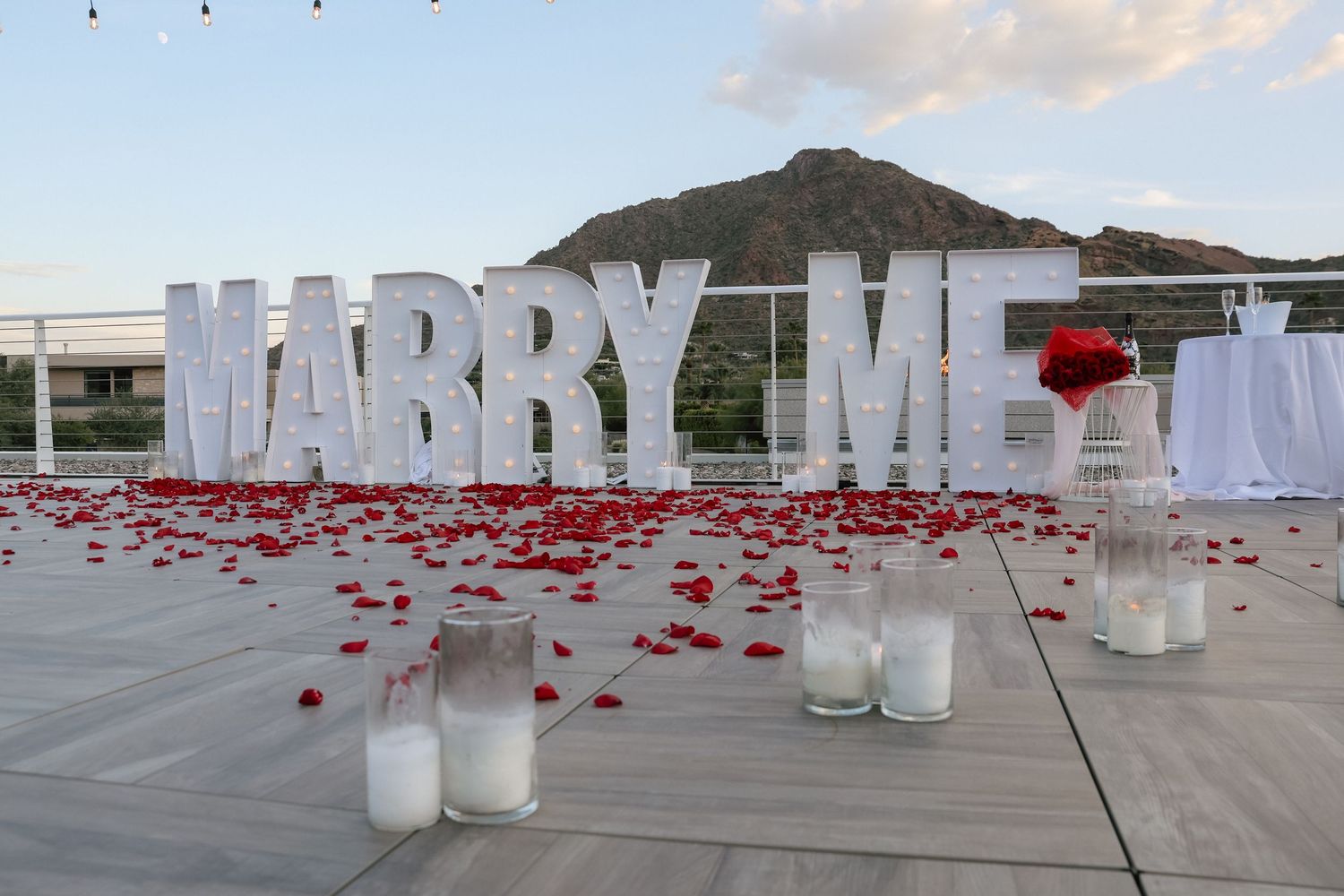Romantic outdoor marriage proposal setup with large MARRY ME letters, red rose petals, and candles on a terrace.