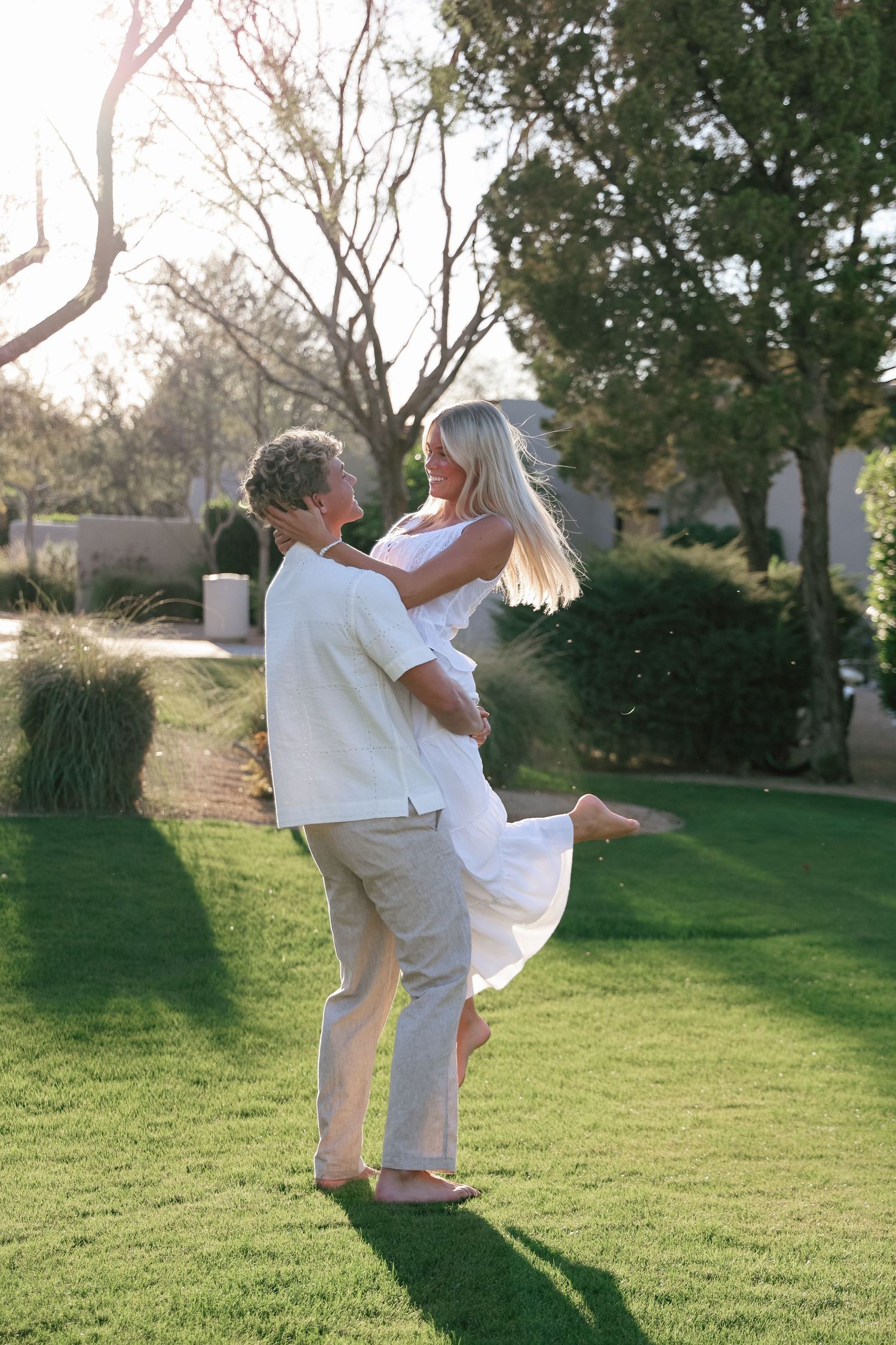 A man lifts a smiling blonde woman in a white dress on a sunny green lawn surrounded by trees.