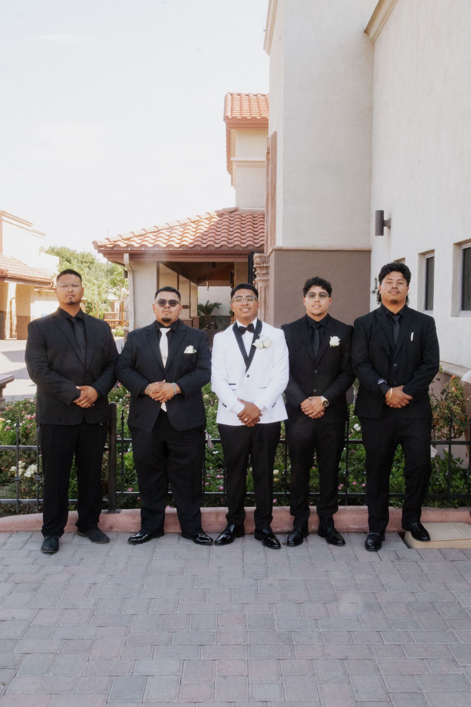 Row of groomsmen in black suits sit together on stone pavement during wedding day.
