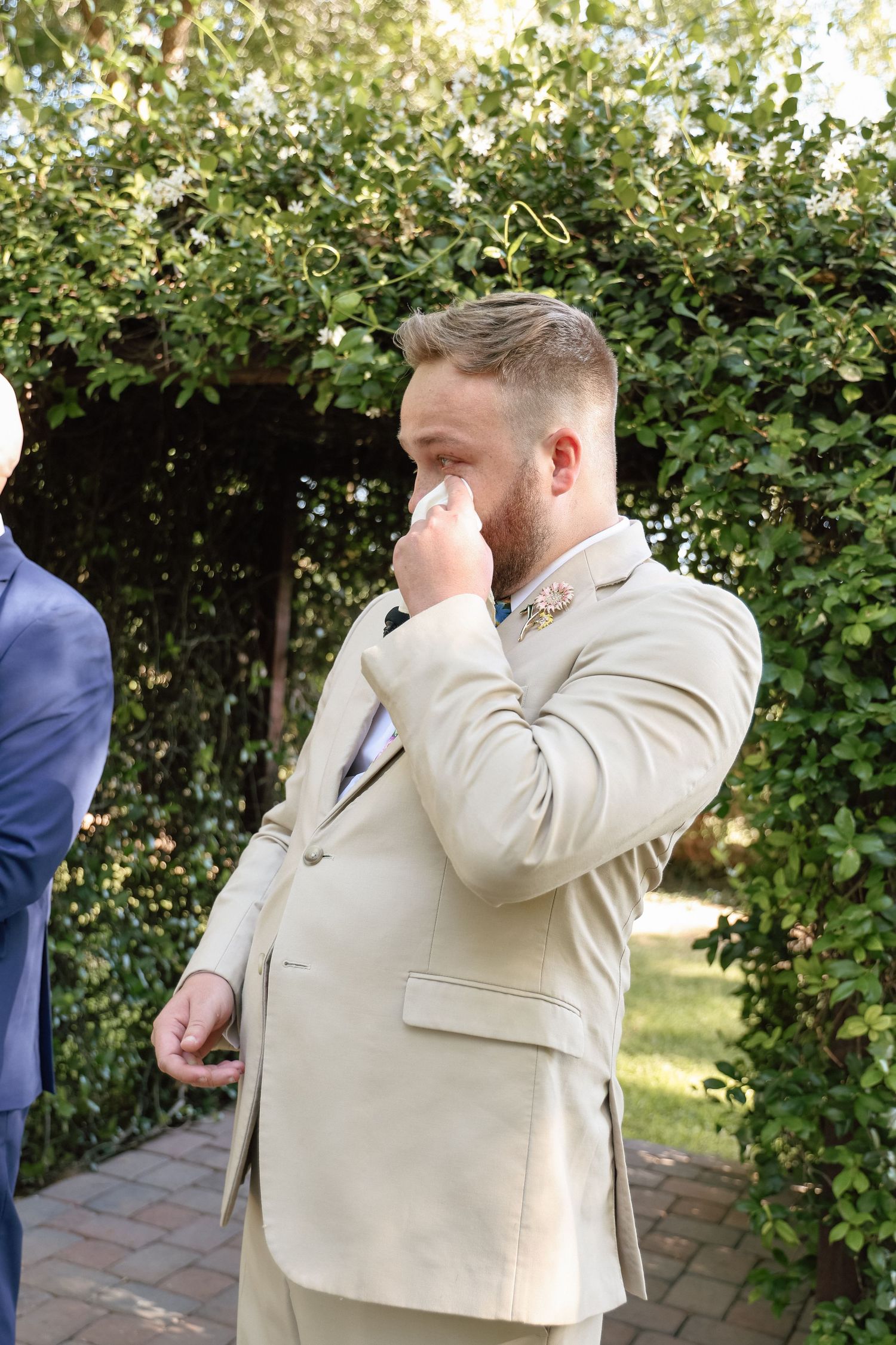 Emotional groom in beige suit wiping tears during outdoor wedding ceremony surrounded by green foliage.