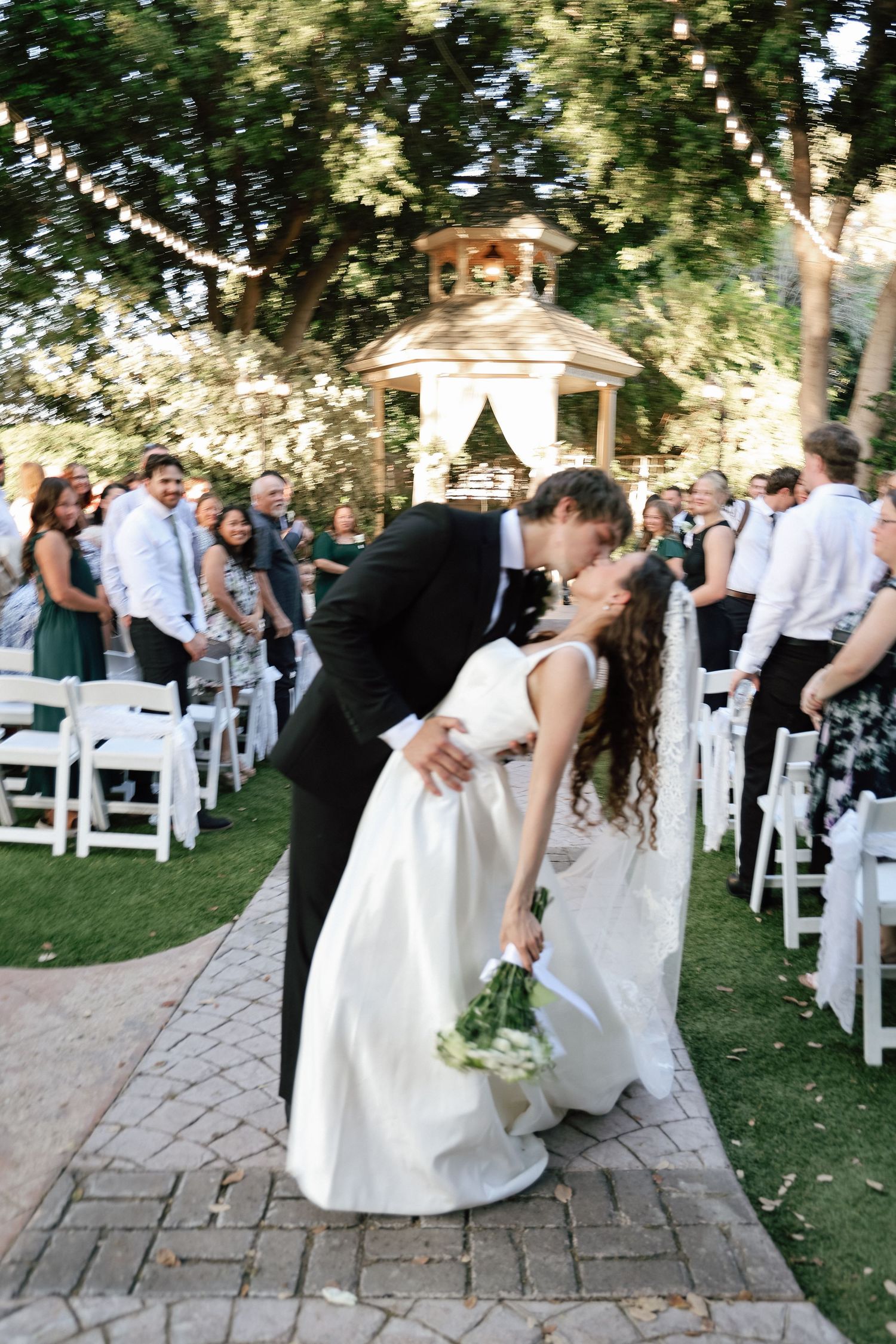 Bride and groom share a romantic dip kiss at outdoor wedding ceremony with white chairs and gazebo backdrop.