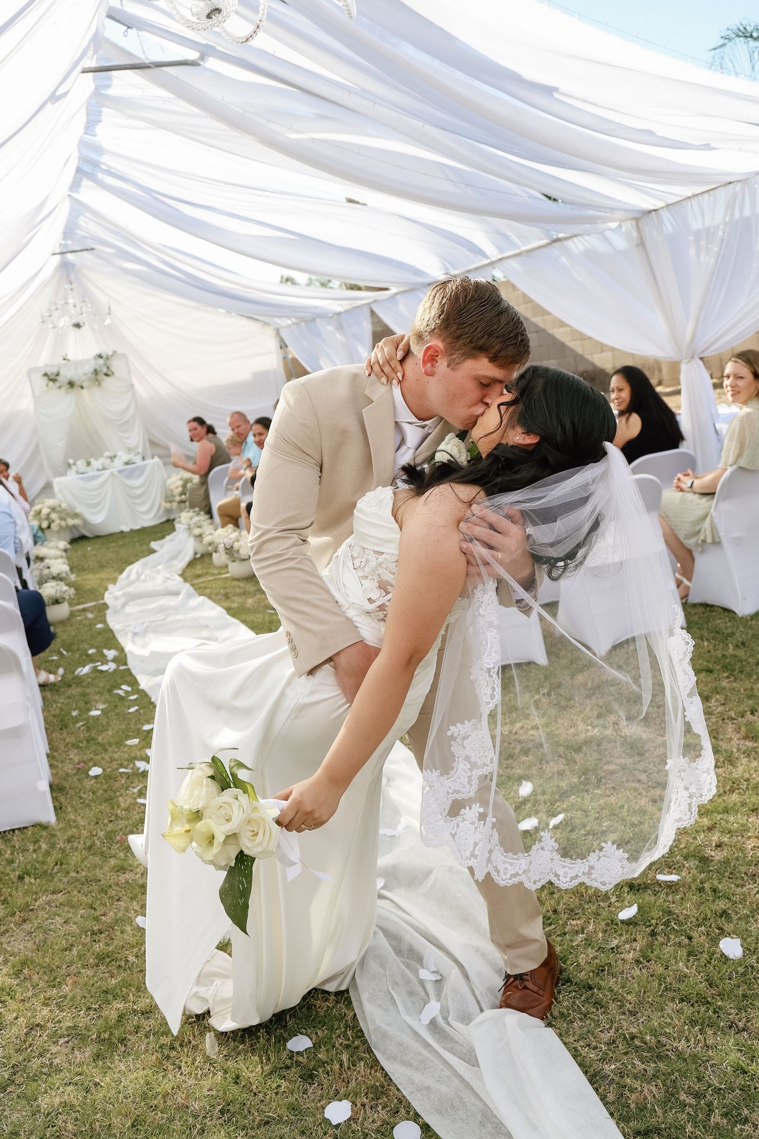 Bride and groom sharing a romantic dip kiss at outdoor wedding ceremony under white draped canopy.