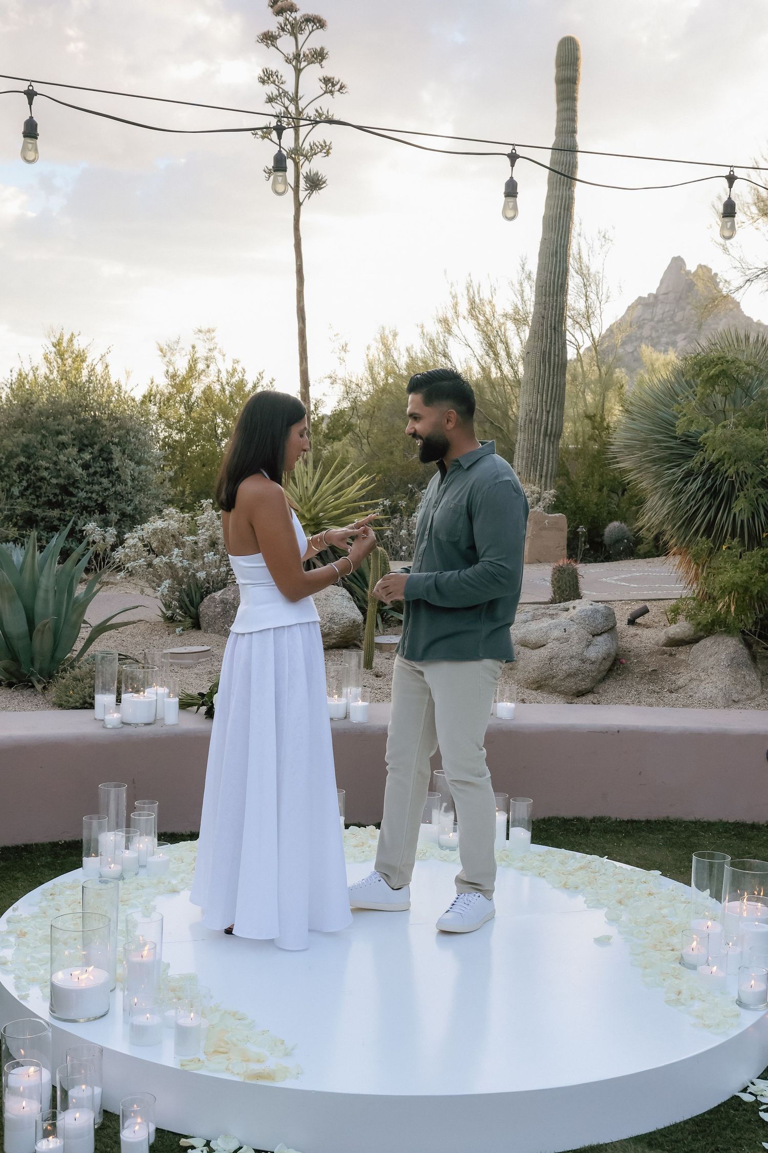 Couple standing on a circular platform surrounded by candles during an outdoor proposal in a desert garden setting.