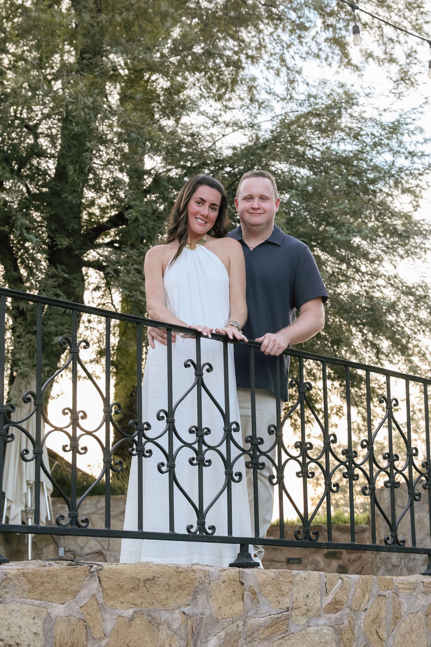 Smiling couple in white dress and polo shirt posing on ornate iron railing with stone steps and trees behind.