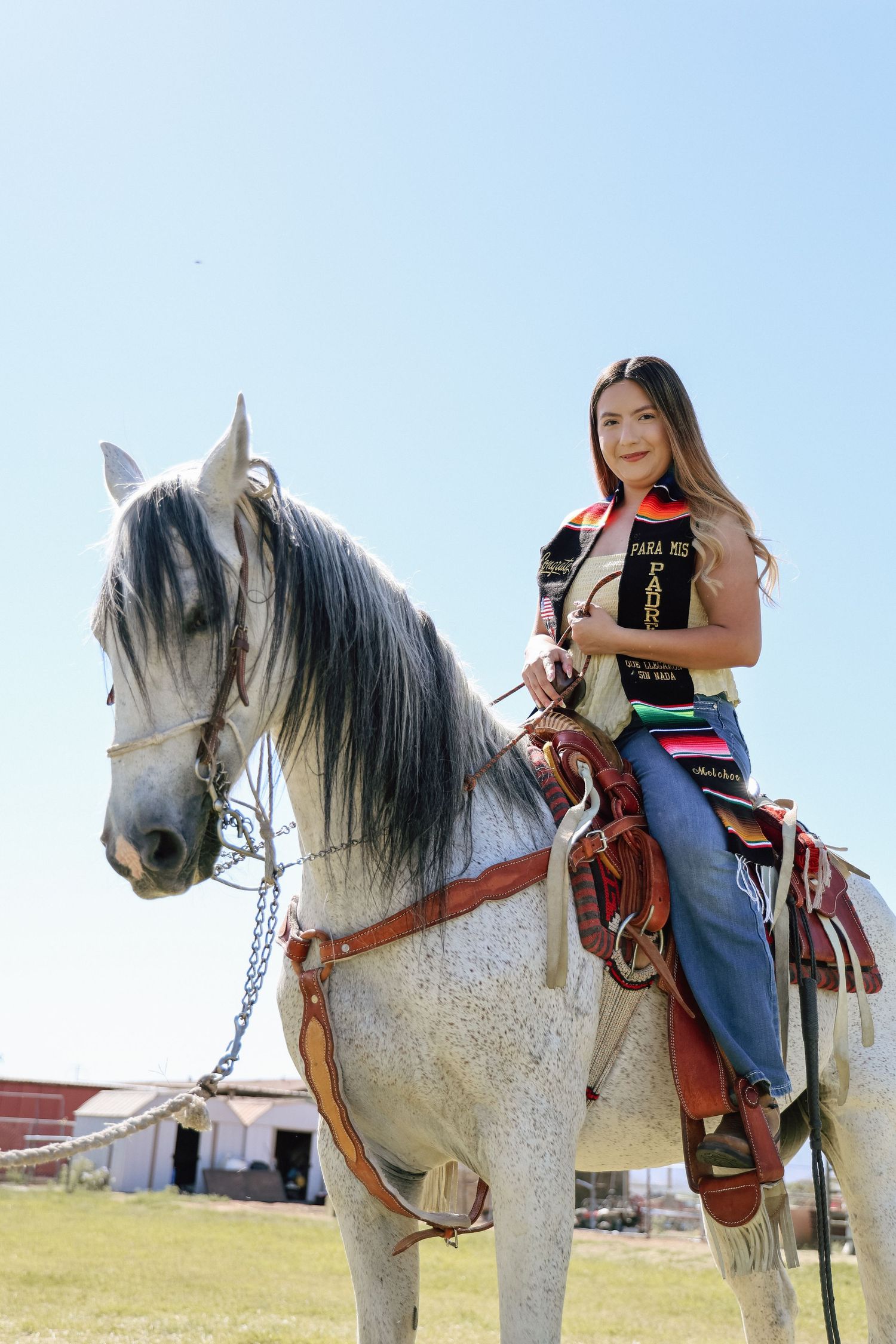 Young woman in traditional attire riding a white horse in an open field under a clear blue sky.