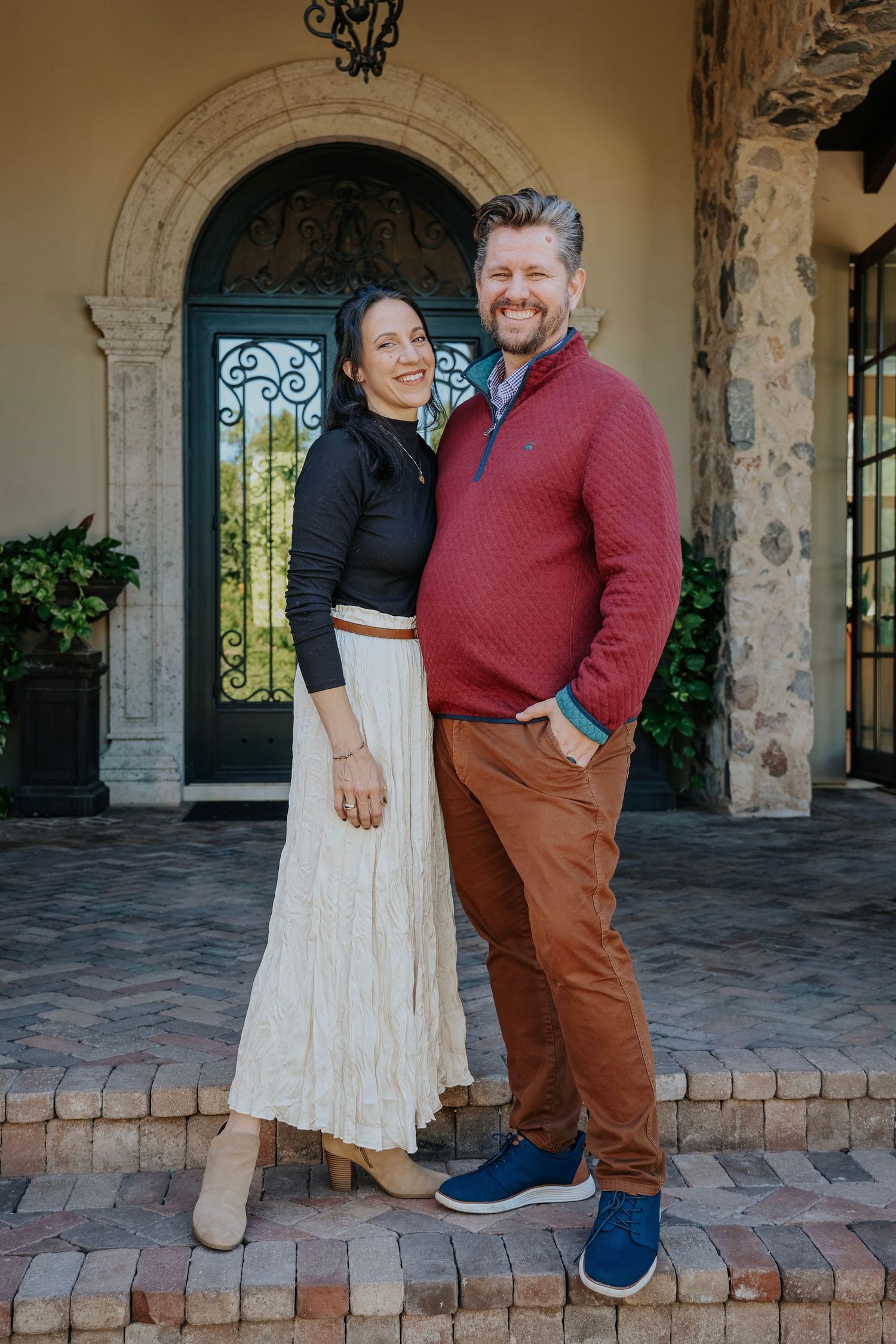 A smiling couple poses together in front of an elegant arched doorway with wrought iron details on brick steps.