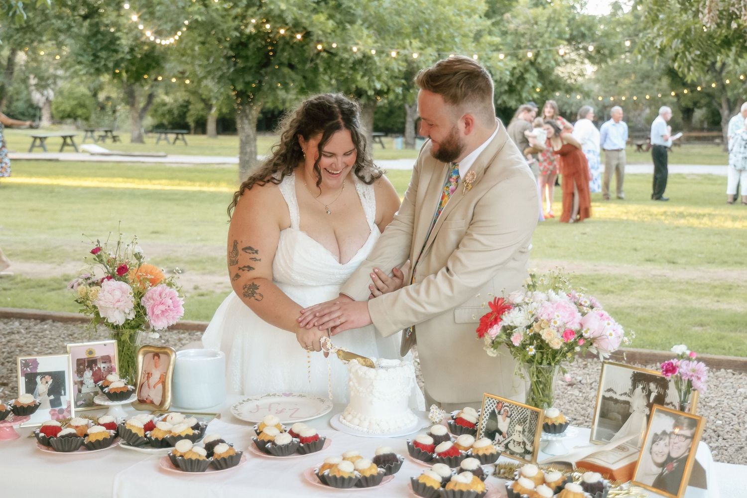Bride and groom cutting wedding cake outdoors at reception table with cupcakes, flowers, and string lights in background.