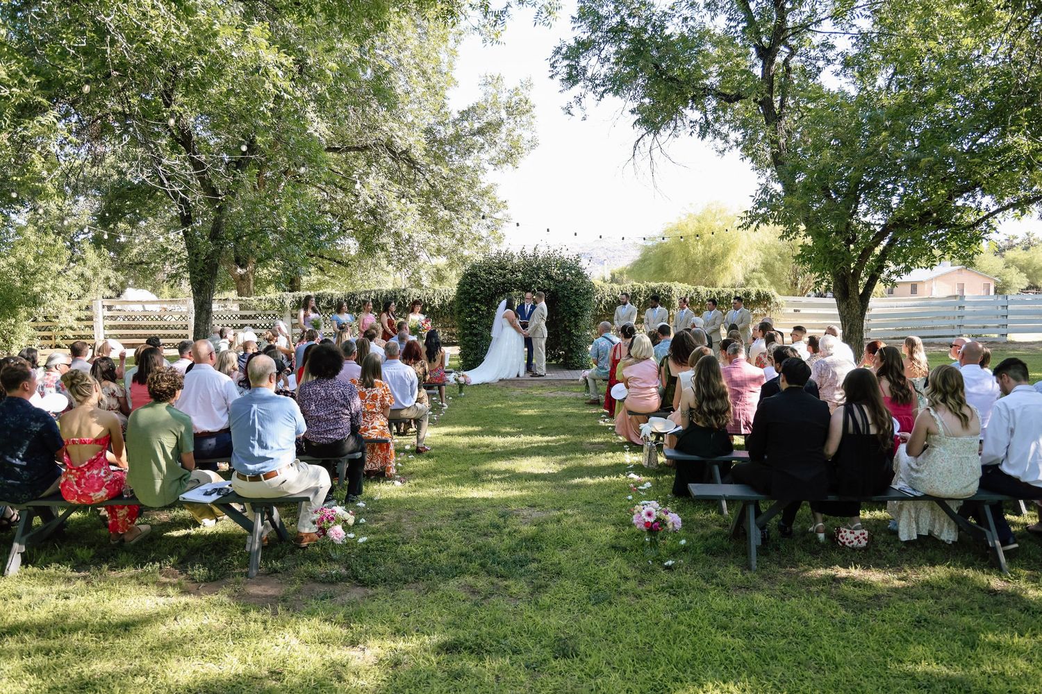 Outdoor wedding ceremony on green lawn with guests seated in rows under shady trees, bride and groom at altar.