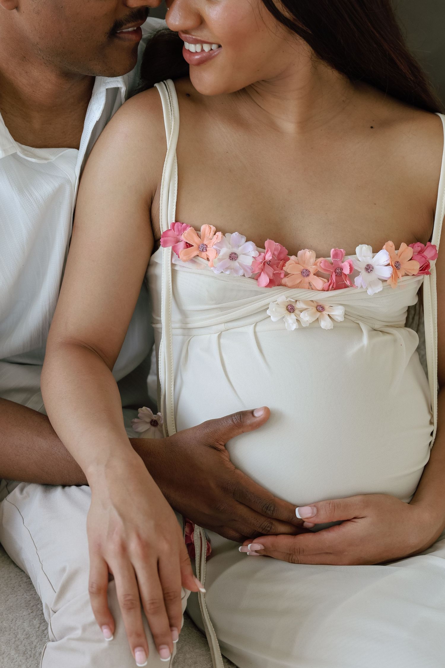 Pregnant woman in white floral dress cradling baby bump with partner beside her smiling.