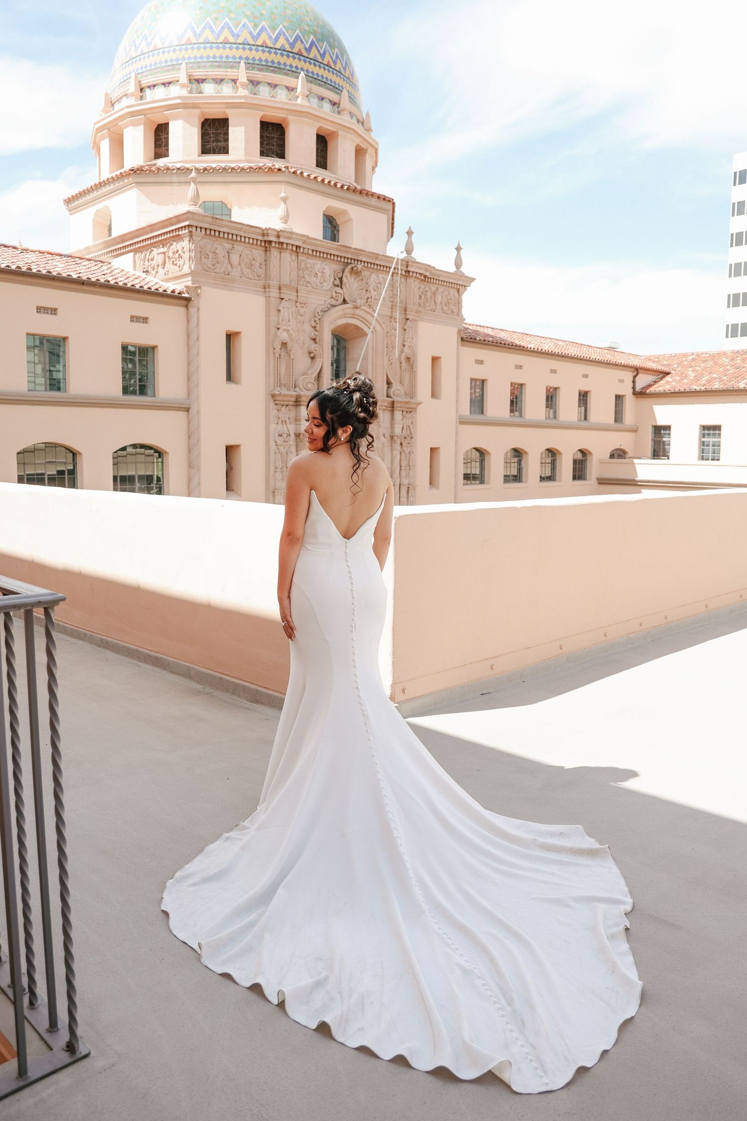 A white wedding dress with a flowing train photographed against a Spanish colonial style building with domed architecture.