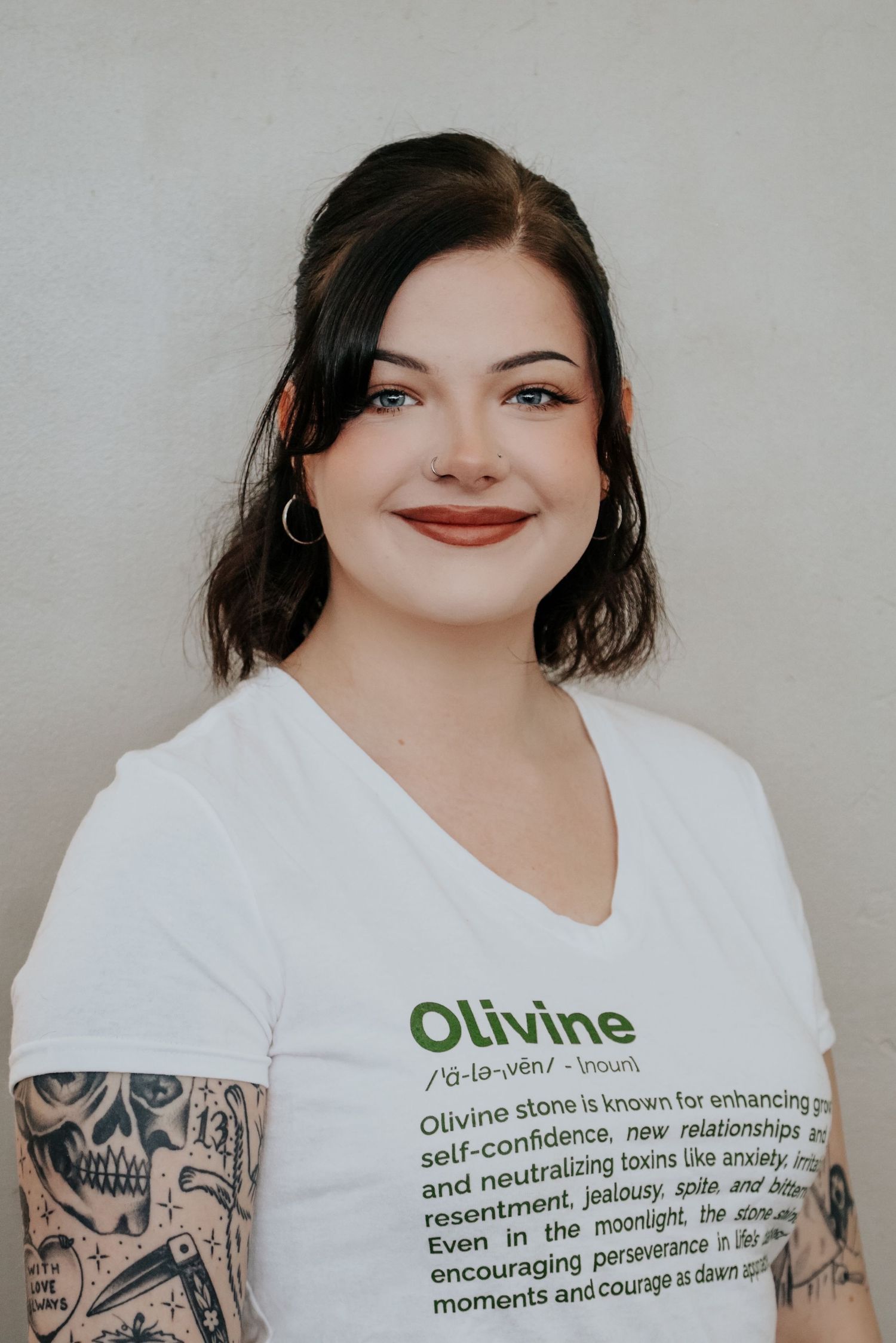 Person with dark hair and tattoos wearing white t-shirt with Olivine stone text.