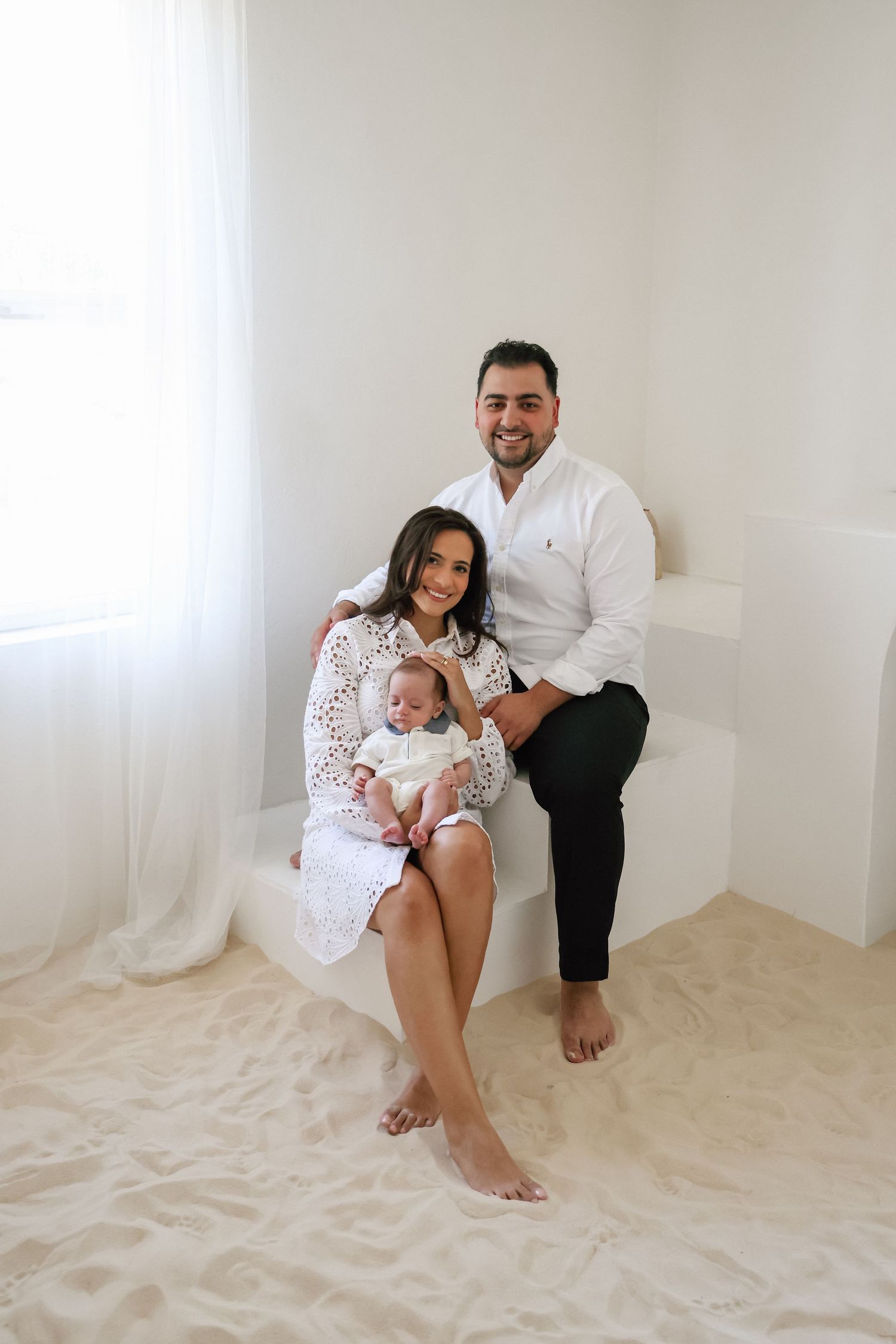 Happy family portrait with parents in white outfits holding newborn baby, seated on sandy white studio floor.