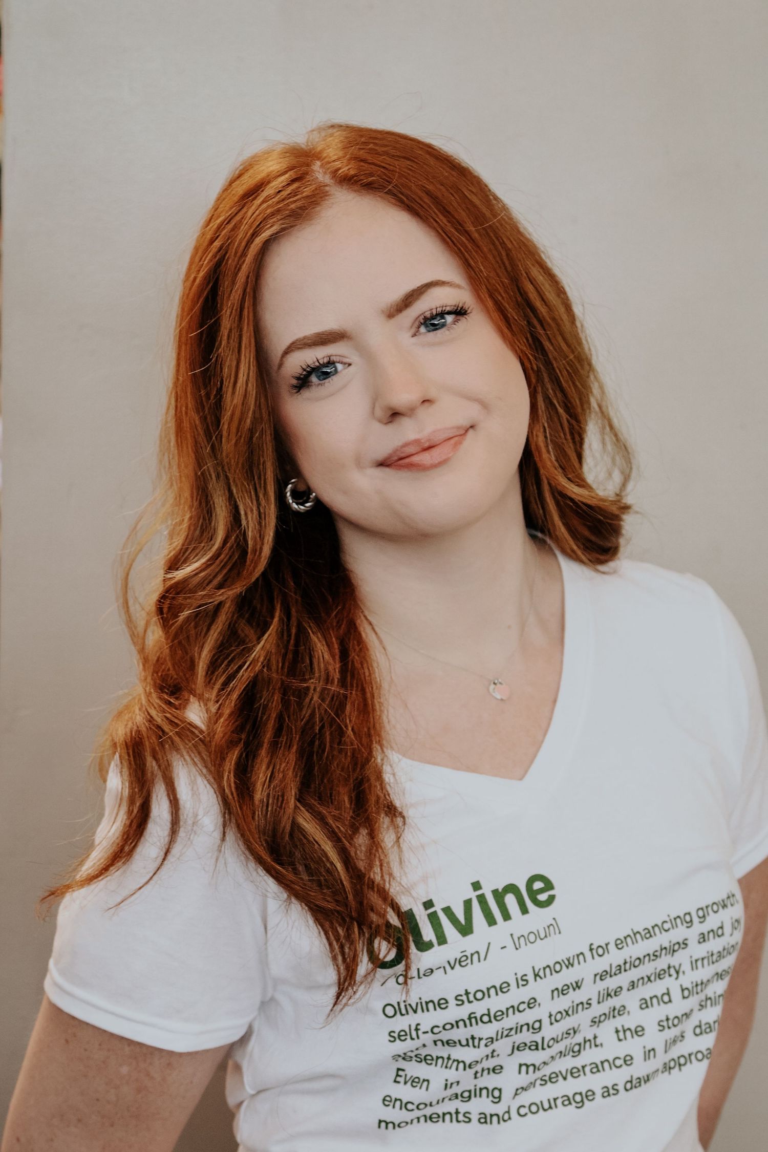 Person with red hair wearing white t-shirt with text about Olivine stone against neutral background.