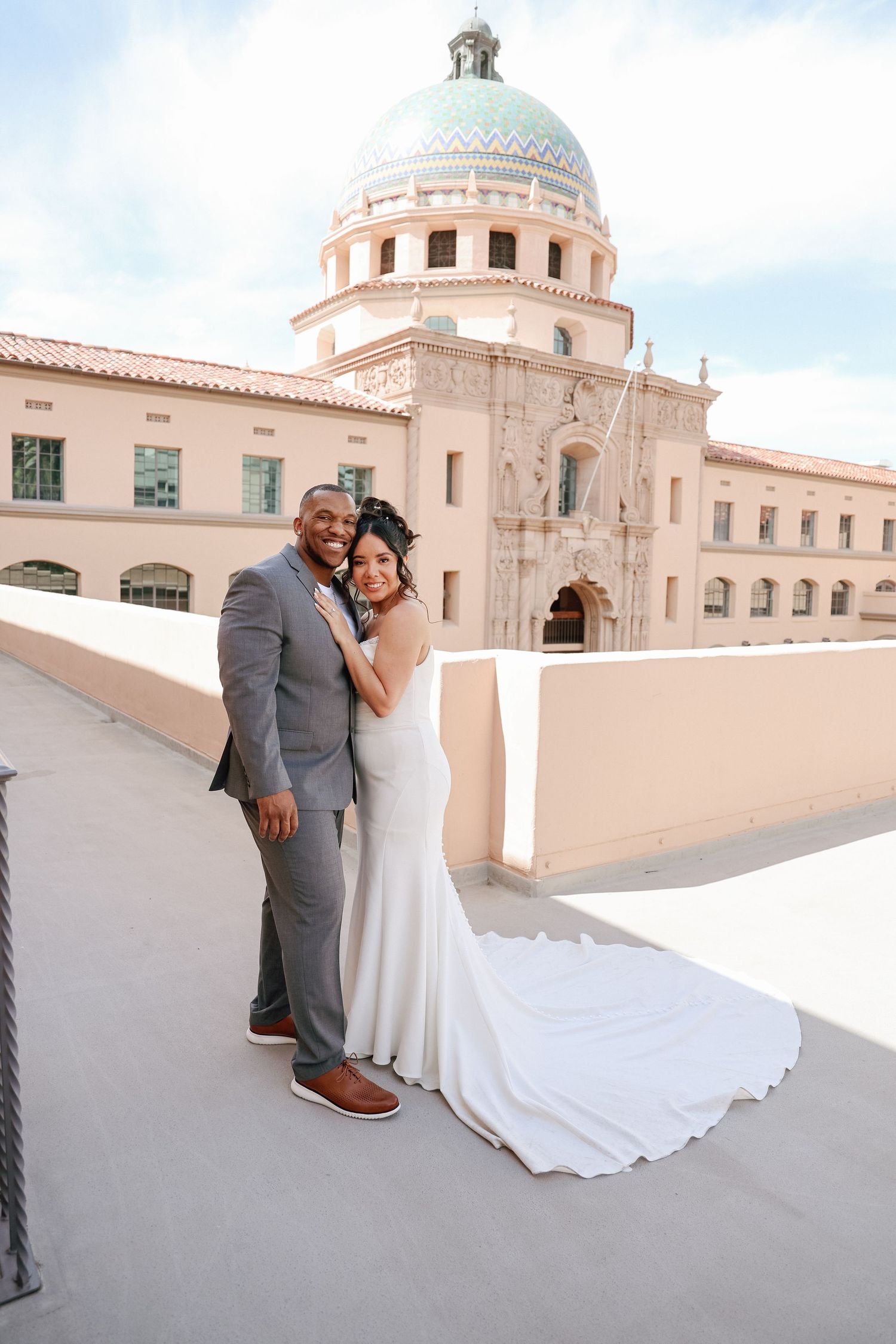 A wedding couple poses in front of a beautiful domed Spanish colonial building with peach-colored walls.