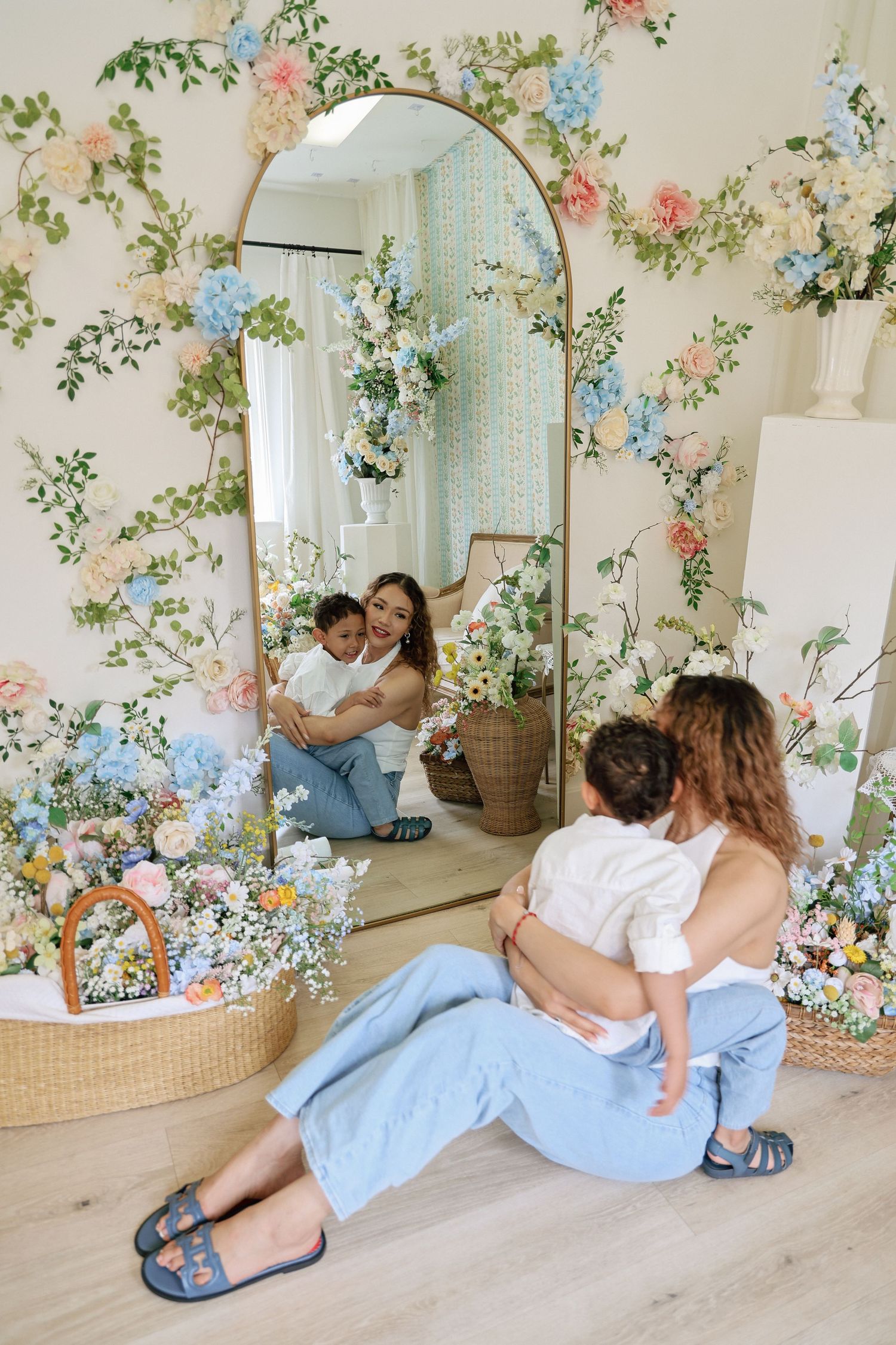 A woman sits on the floor holding a baby, reflected in an arched mirror, surrounded by floral arrangements and pastel decor.