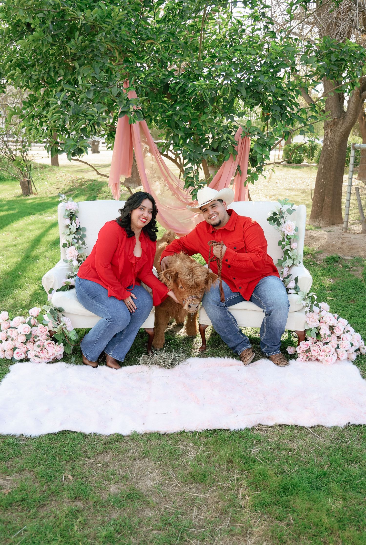 Couple in matching red hoodies sitting on a white sofa with a baby cow, surrounded by pink flowers outdoors.