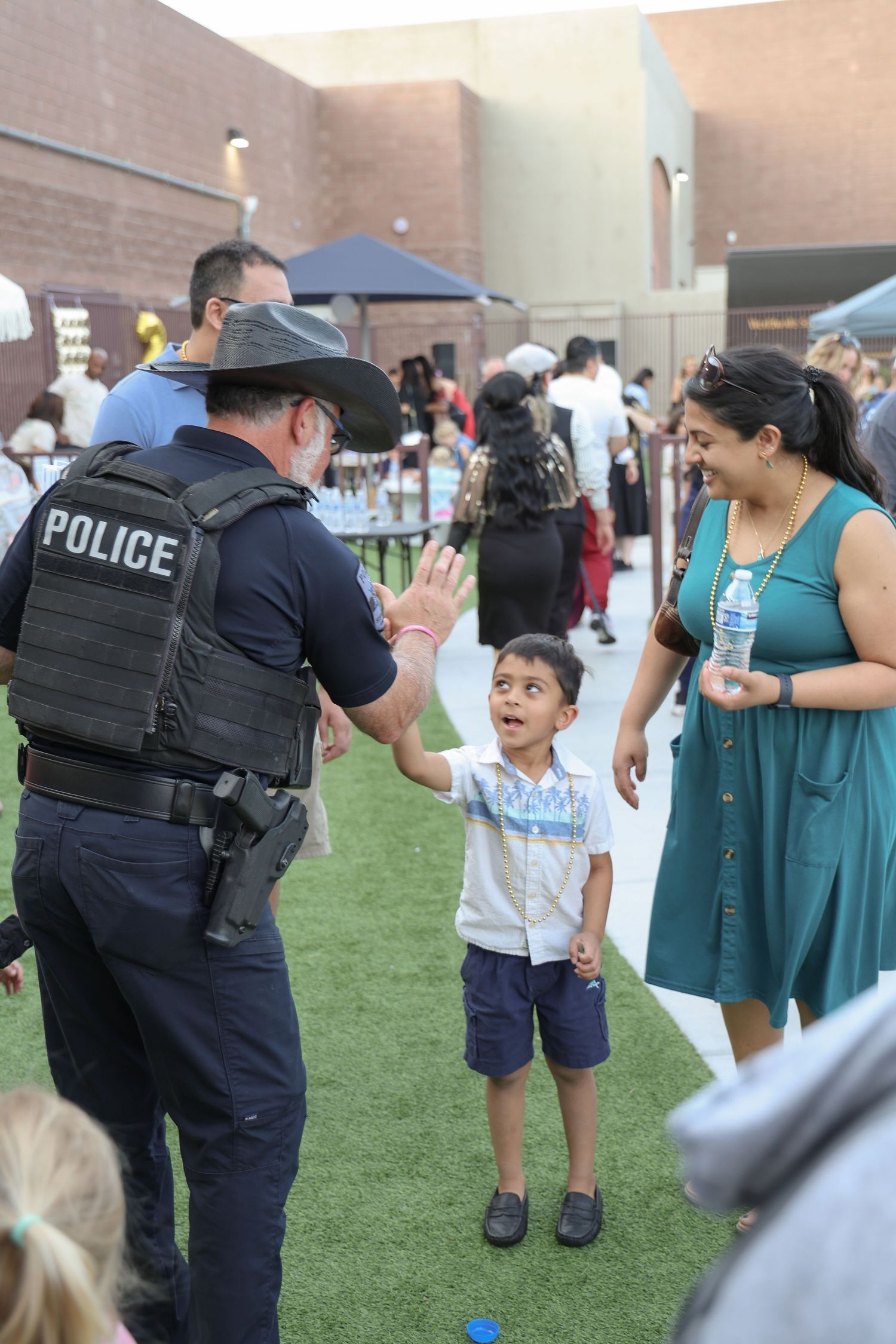 Police officer in tactical vest interacts with children and families at community outreach event on grass lawn.