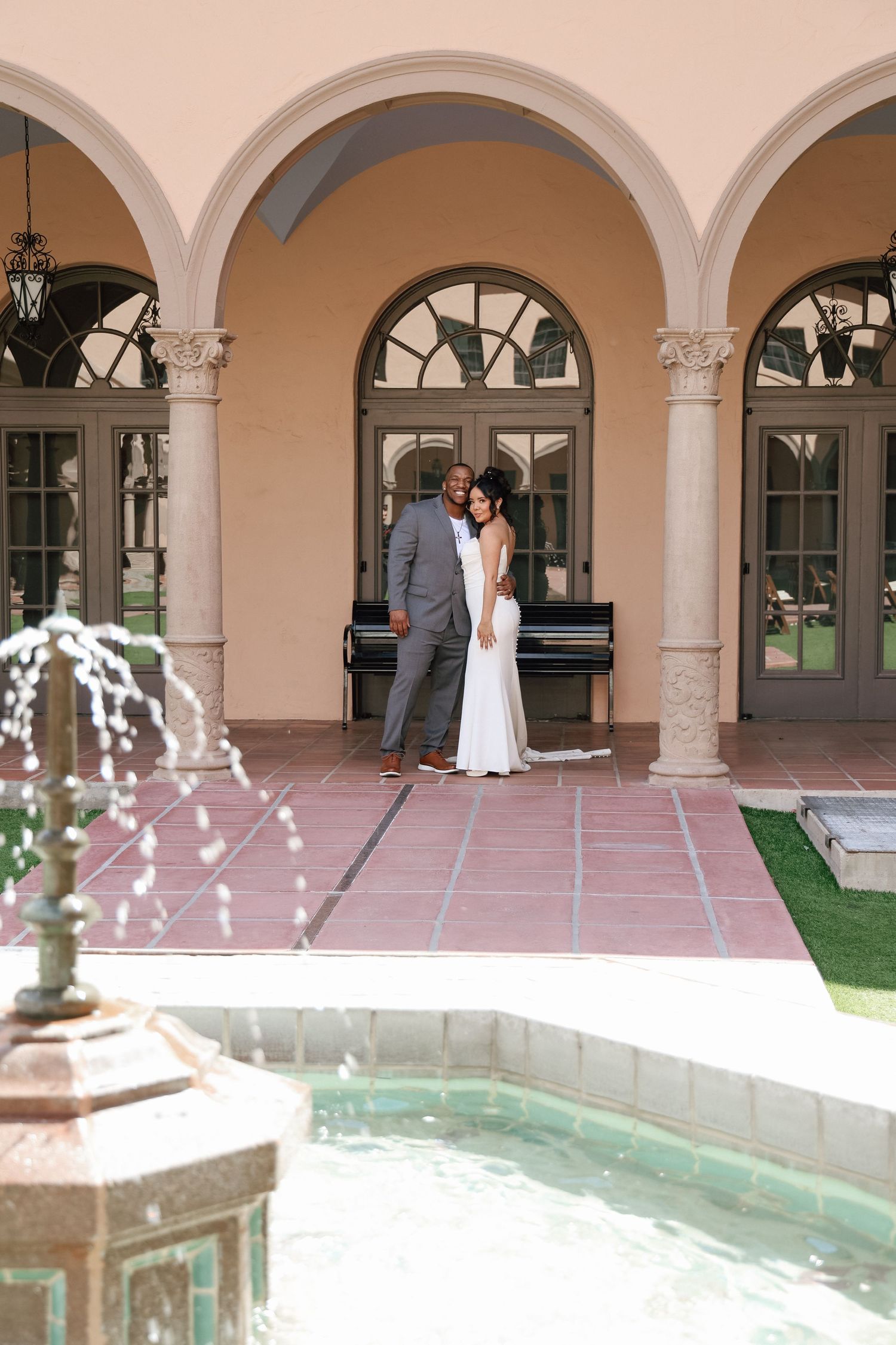 A couple poses under arched columns near a fountain in a Spanish colonial style courtyard.