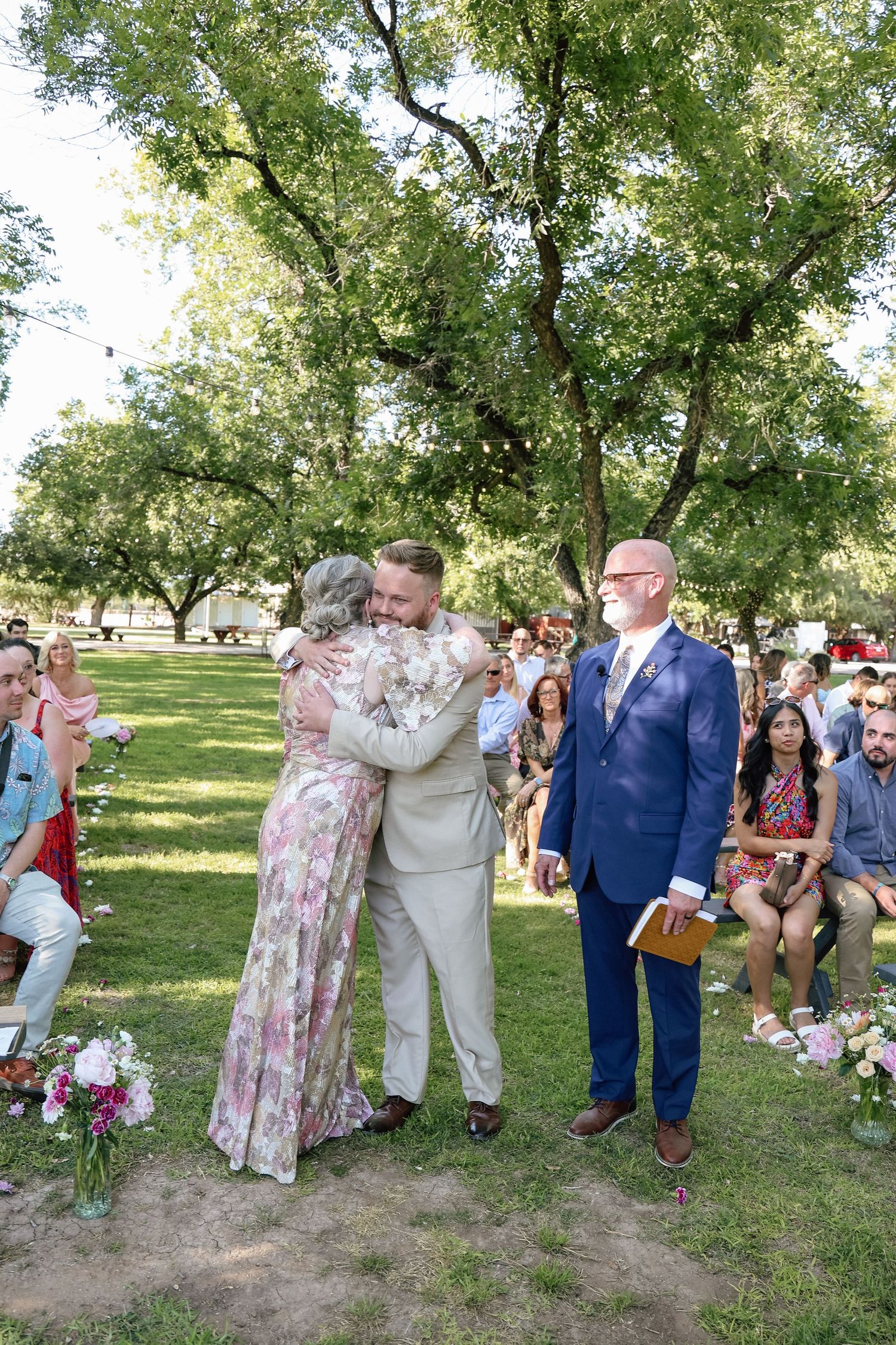 Groom hugging a woman in floral dress during outdoor wedding ceremony while officiant and guests watch.