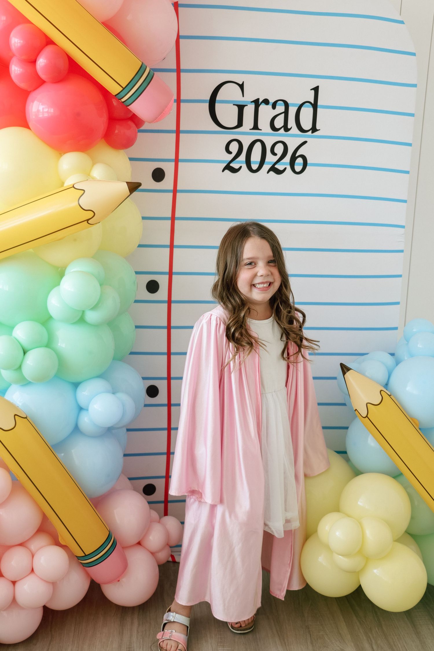 Young girl in pink graduation gown smiling in front of a Grad 2026 backdrop with colorful balloons.