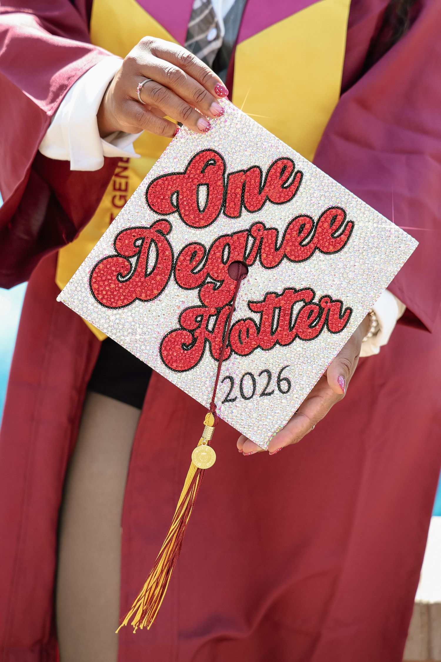 Graduation cap decorated with glitter text reading One Degree Hotter 2026, held by graduate in maroon gown.