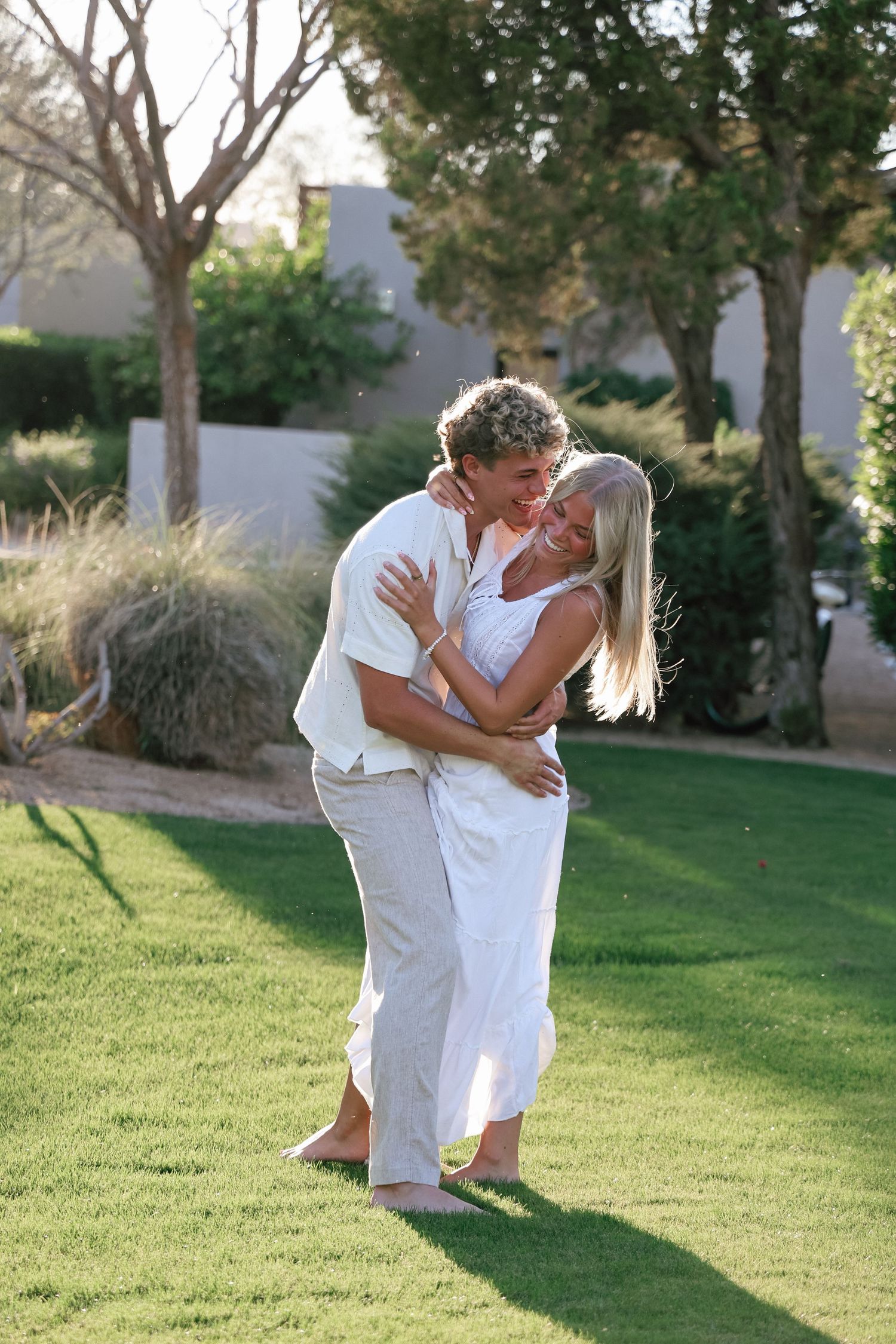 Two people in white clothing share intimate dance moves on a sunny lawn surrounded by trees.