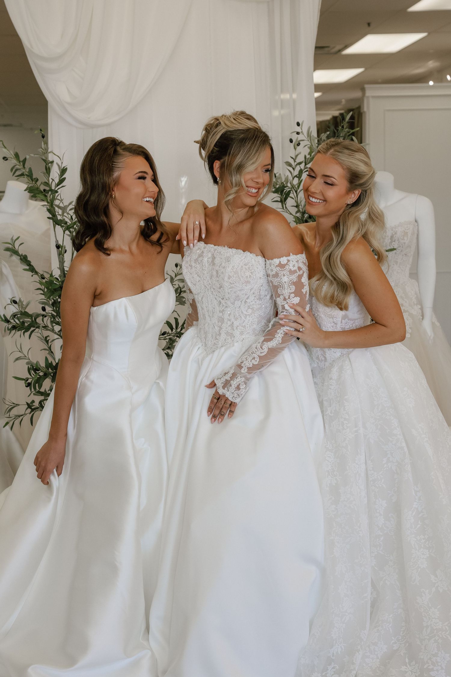 Three women smiling in elegant white wedding gowns inside a bridal boutique.