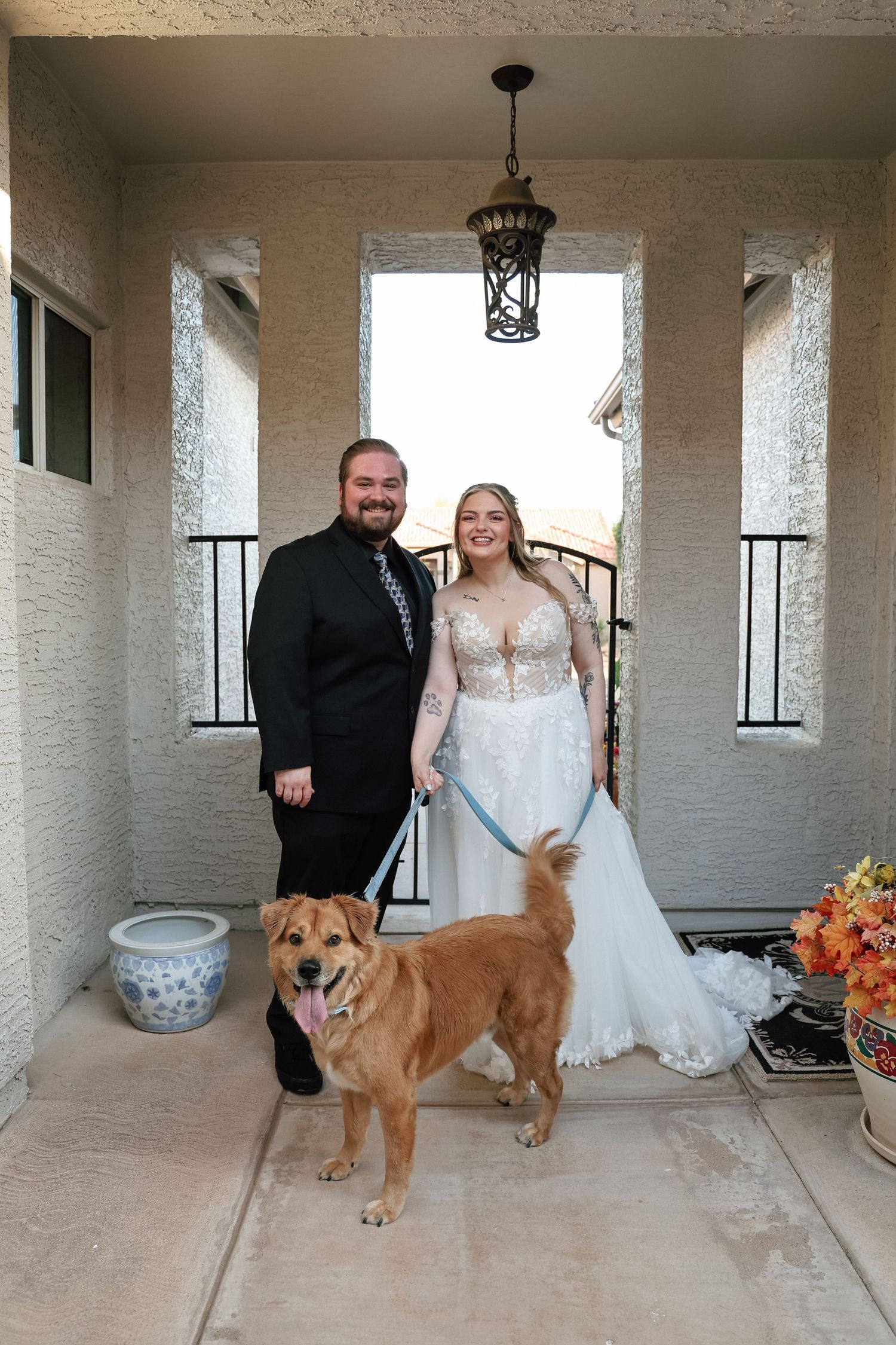 Newlywed couple in wedding attire posing with golden retriever dog at decorated home entrance.