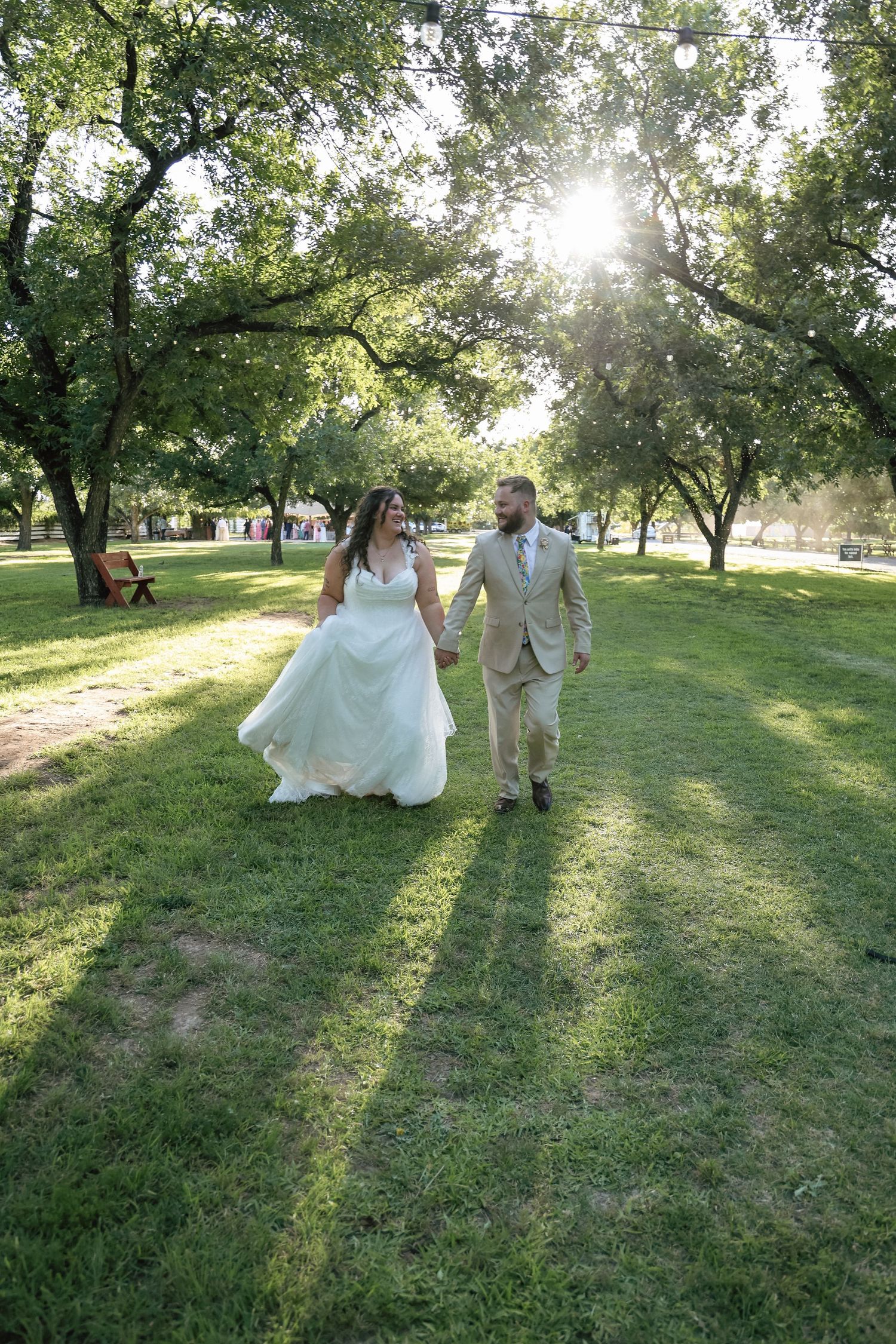 Bride and groom walking hand in hand through sunny green park on their wedding day.