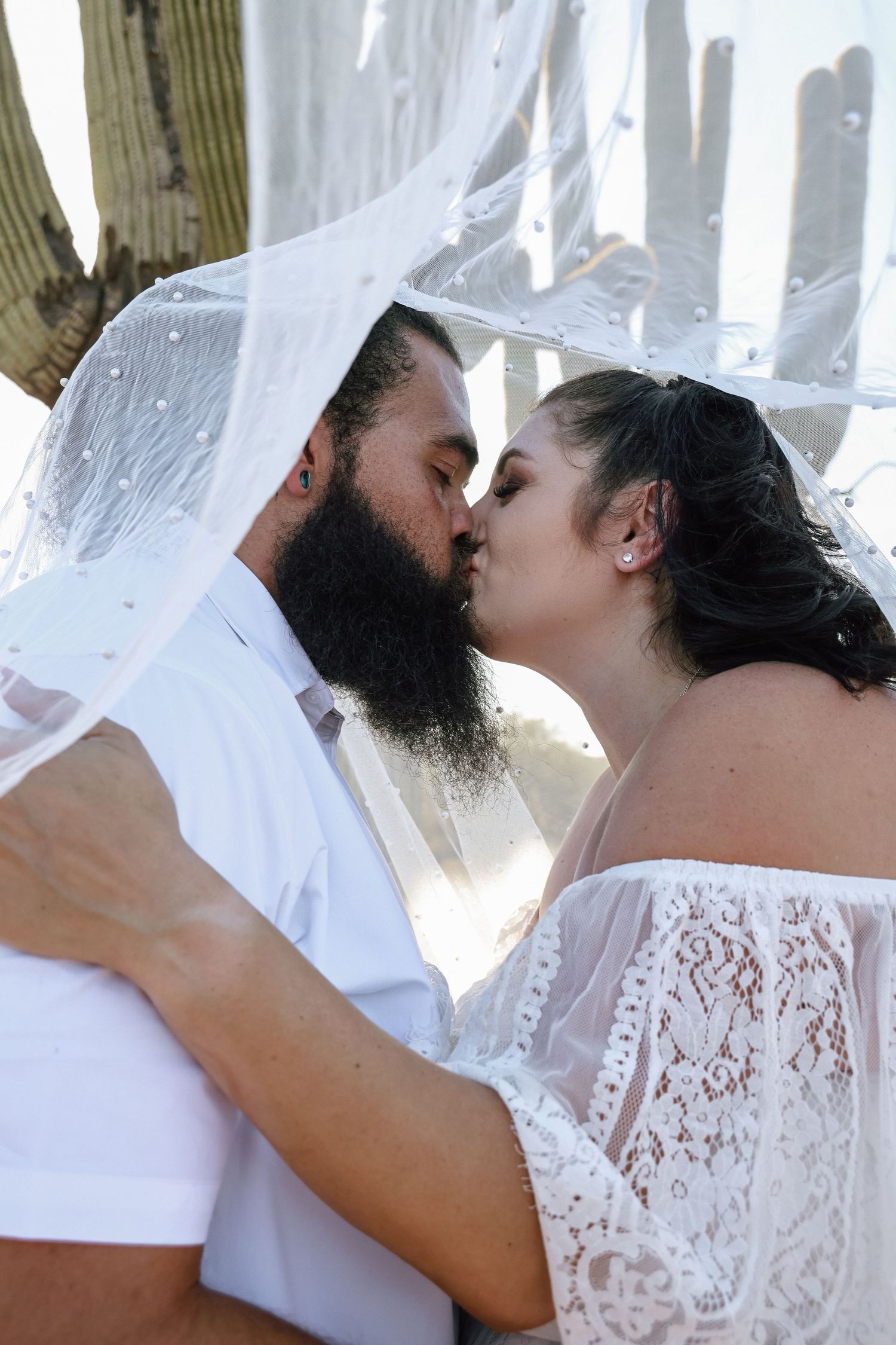 Bride and groom sharing a romantic kiss under a white veil at an outdoor wedding ceremony.