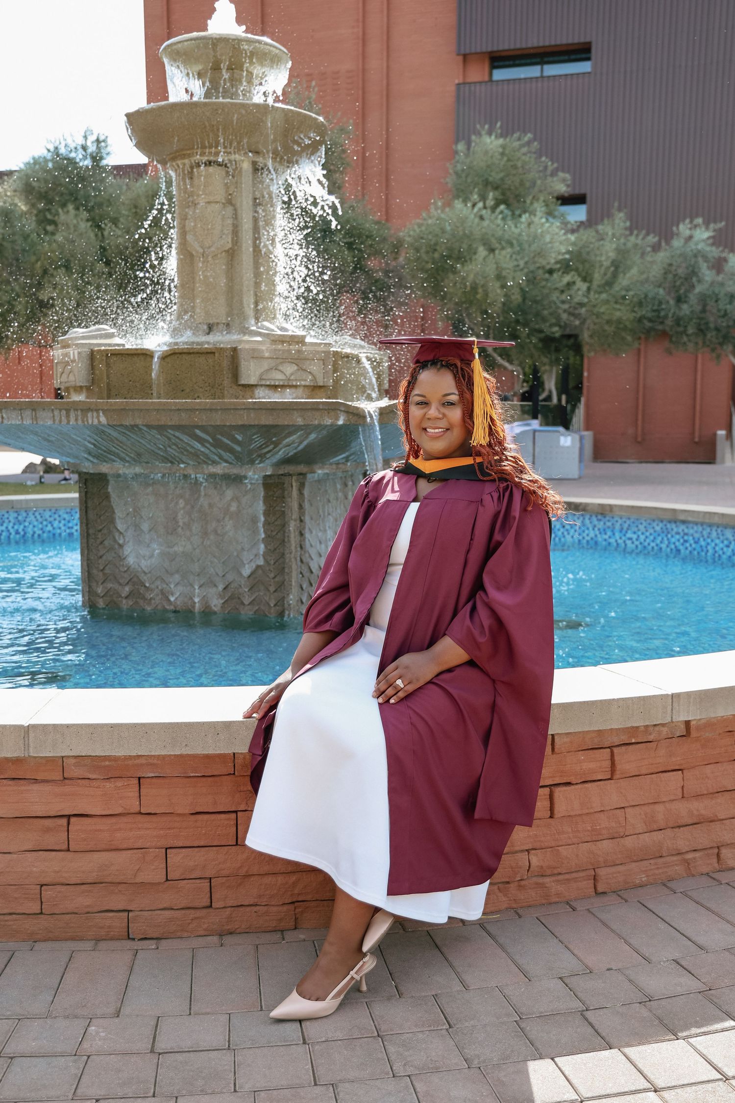 Graduate in maroon cap and gown poses by ornate fountain on college campus on graduation day.