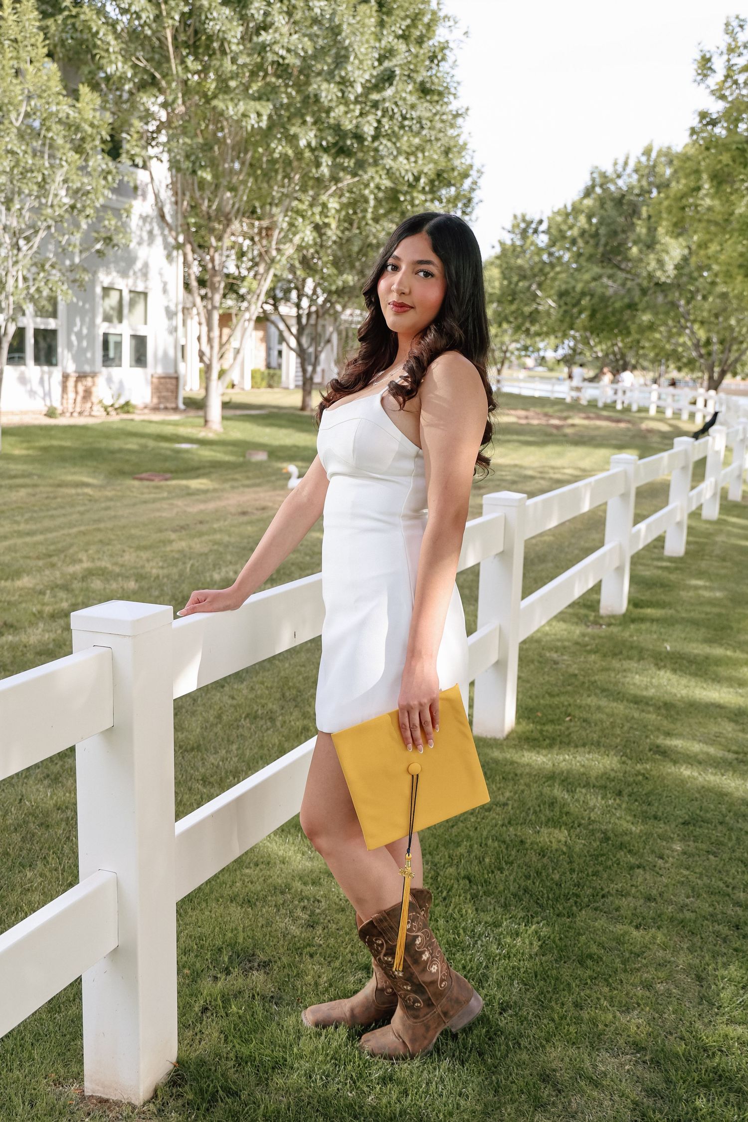 Young woman in white dress and brown cowboy boots holding yellow graduation cap by white fence.
