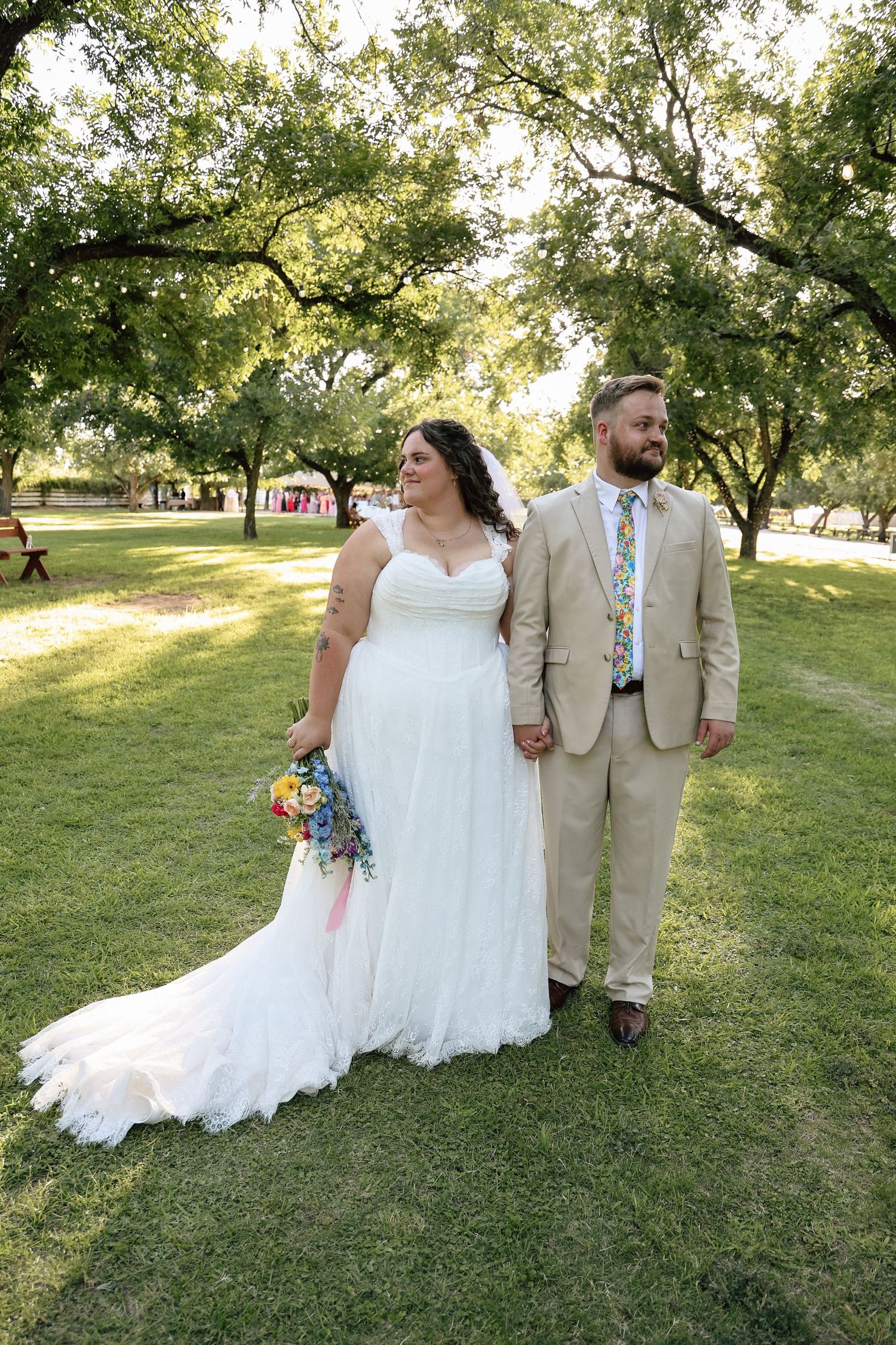 Bride in white gown and groom in tan suit hold hands while posing together in a lush green park.